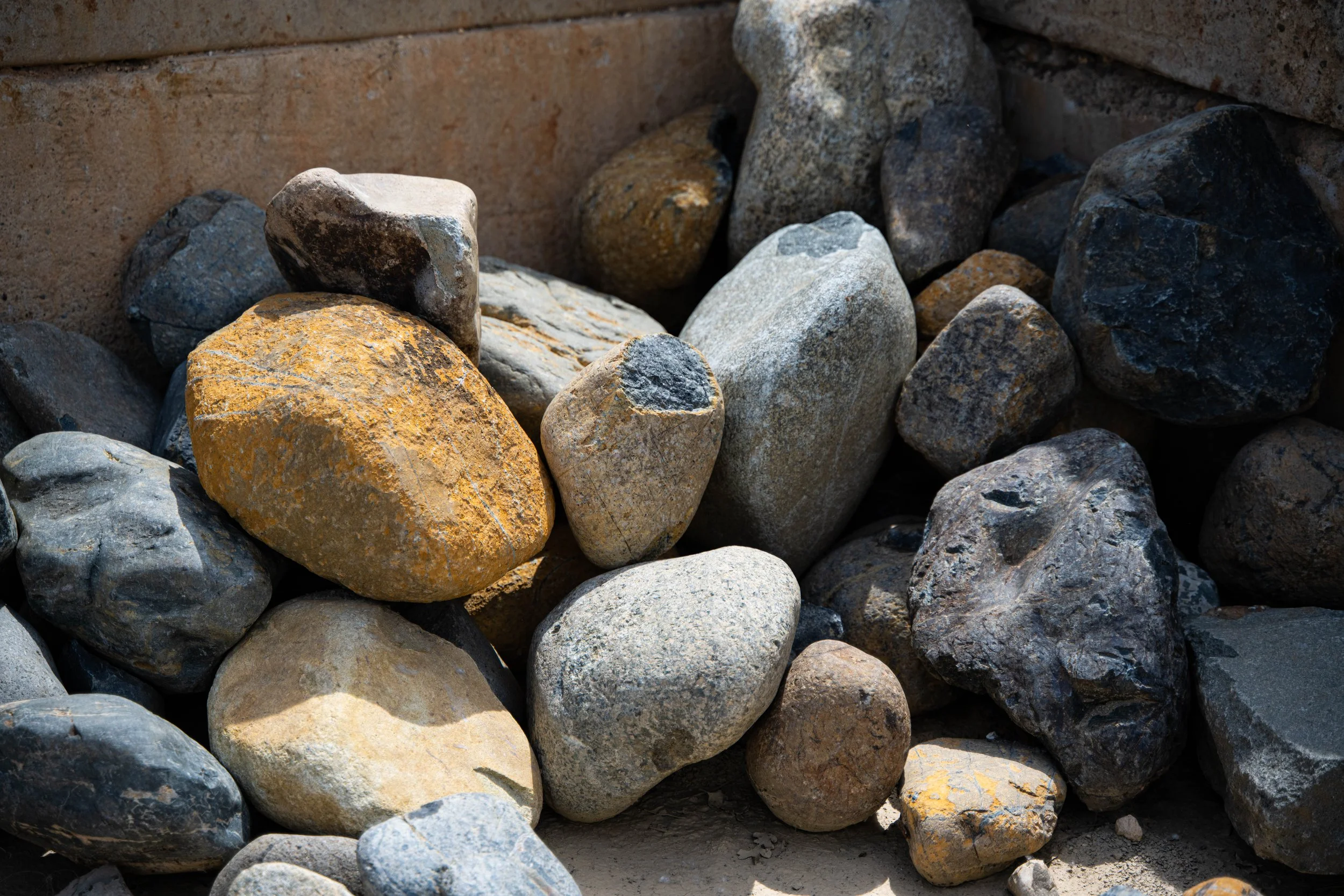 A pile of multicolored rocks and stones ranging in size, with some showing yellow lichen, set against a background of earth and some bricks.