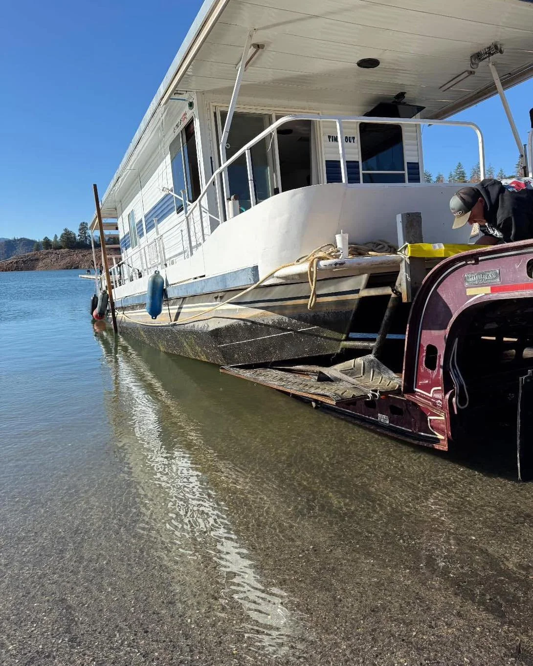 A houseboat is docked at the shoreline with a person working on it near a trailer. The houseboat has an overhanging roof and windows, with a tan exterior and blue fenders hanging on the side. The boat is floating on calm water with a clear blue sky overhead.