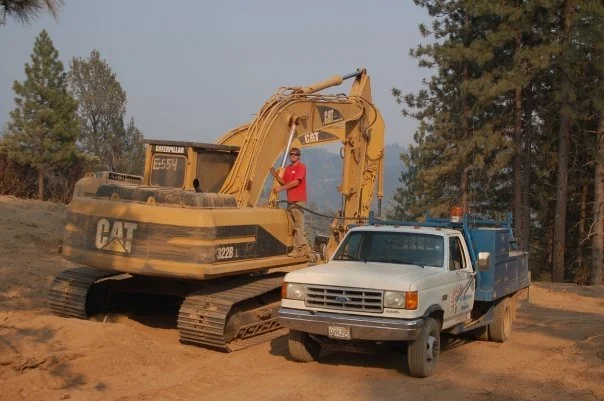 A person stands on a large yellow Caterpillar excavator next to a blue work truck on a dirt surface, with trees in the background.