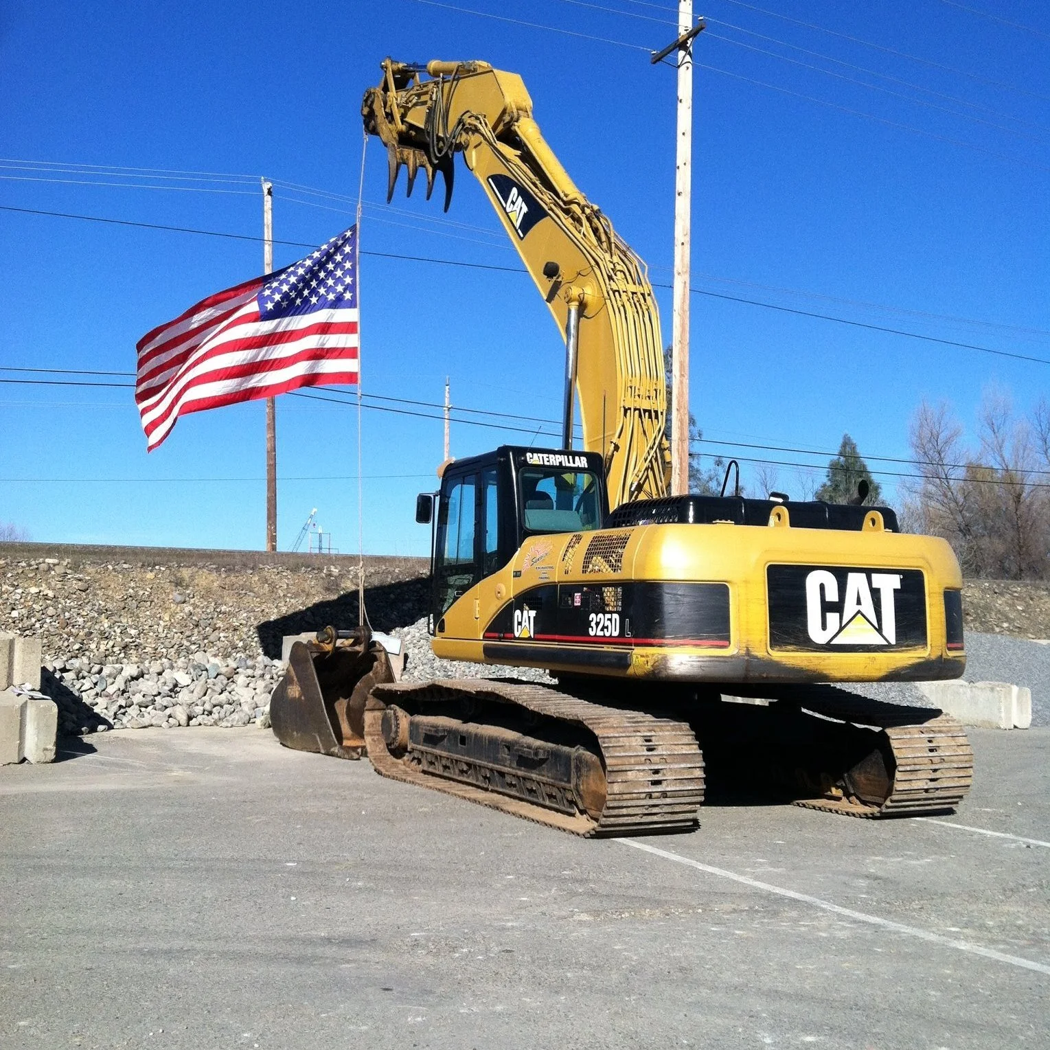 A yellow CAT excavator with a black and white logo, sitting on a paved lot with a gravel embankment in the background, next to an American flag on a flagpole against a clear blue sky.