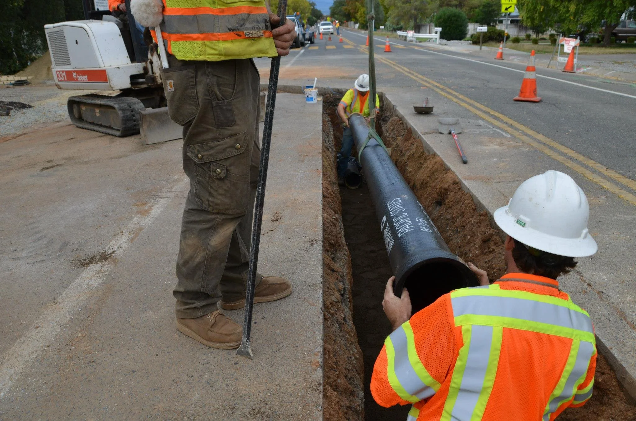 Workers install a large black pipe into a trench on a street, with traffic cones directing traffic and machinery nearby.