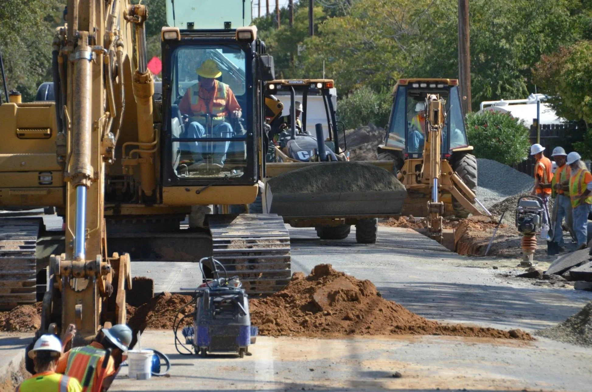 Construction workers using heavy machinery for roadwork; one worker is operating a large excavator, and others are working on the ground with safety gear.