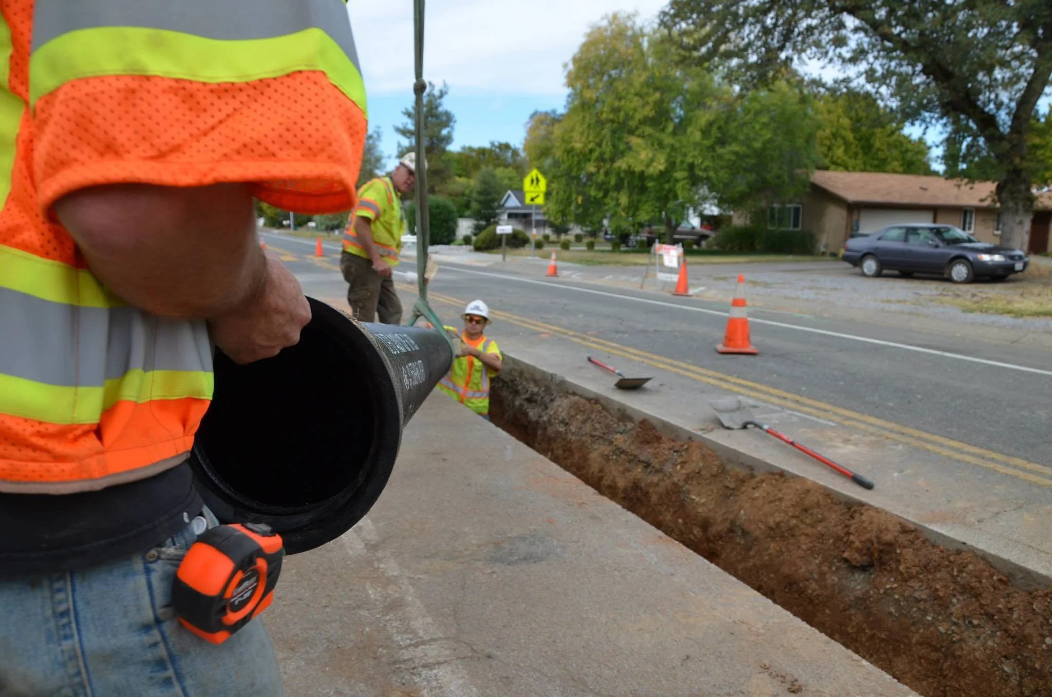 Construction workers in high-visibility vests working on a road with orange cones and digging equipment on the street, with residential houses and trees in the background.