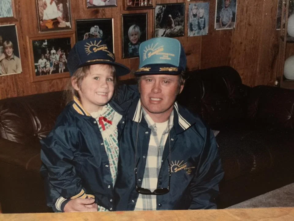 A man and a young girl are sitting on a dark brown leather couch in a room with wood-paneled walls decorated with framed photographs. They are both wearing matching blue caps and jackets with a logo that reads 'Sunshine'. The girl is smiling, and the man appears to be making a surprised or confused facial expression.