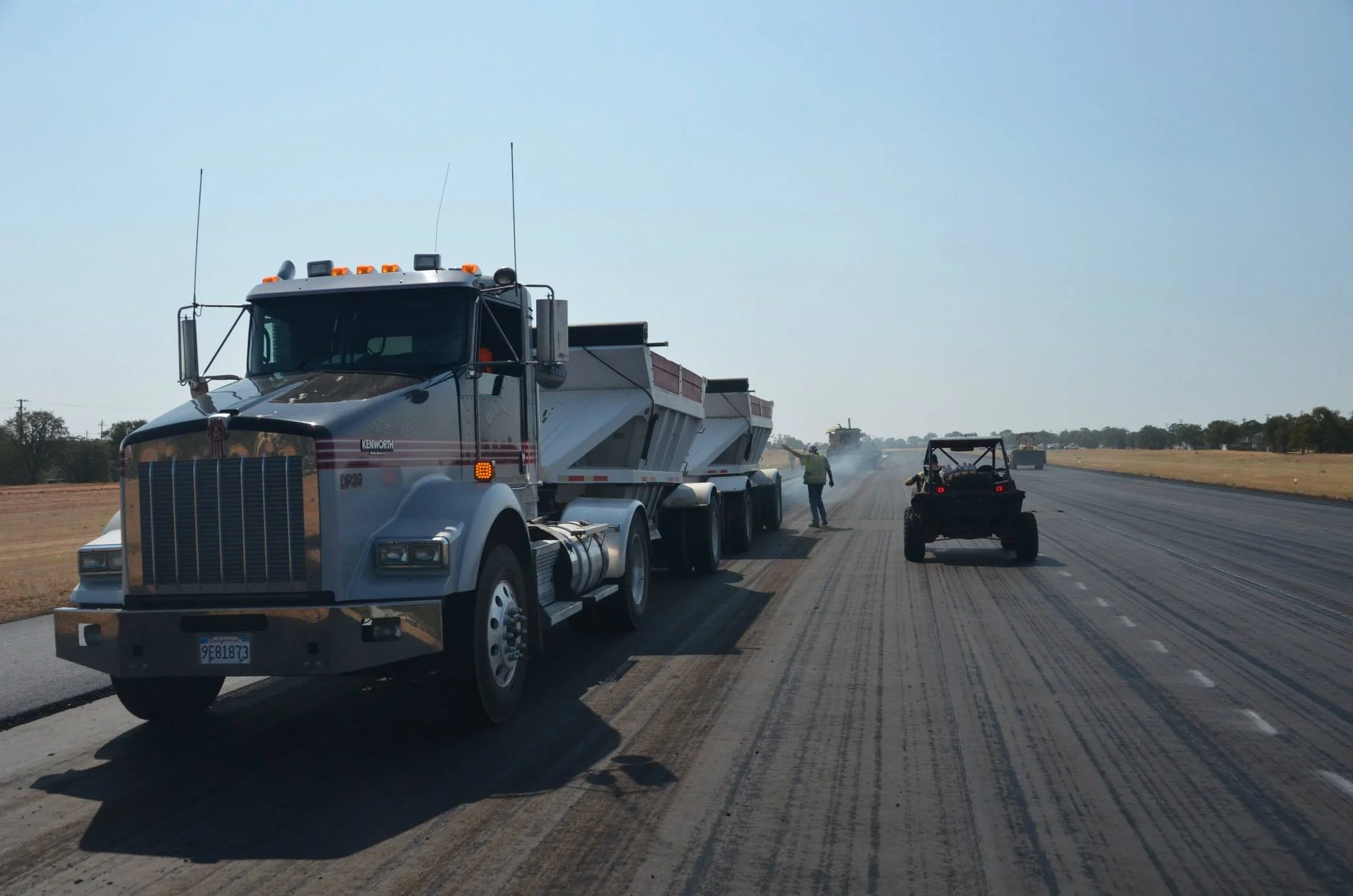 A large Kenworth truck with a white and gray cab and red accents is working on paving a road. There are construction workers and a small roller compactor behind it, with dirt and gravel on the road surface under clear blue skies.