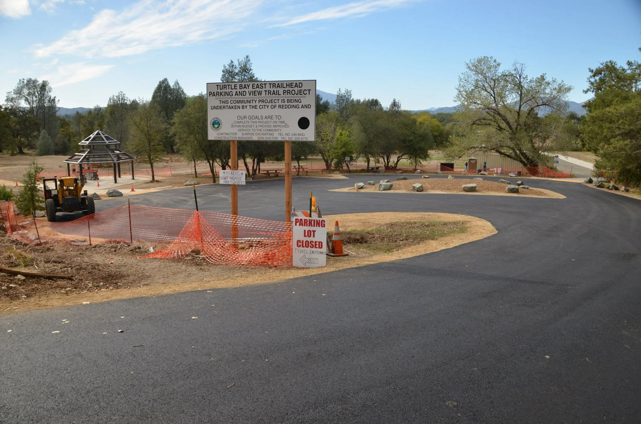 A parking lot is closed for a trailhead construction project, with a sign indicating the project and warning that the lot is closed. The area has trees, rocks, and construction equipment, with a scenic background of hills and a partly cloudy sky.