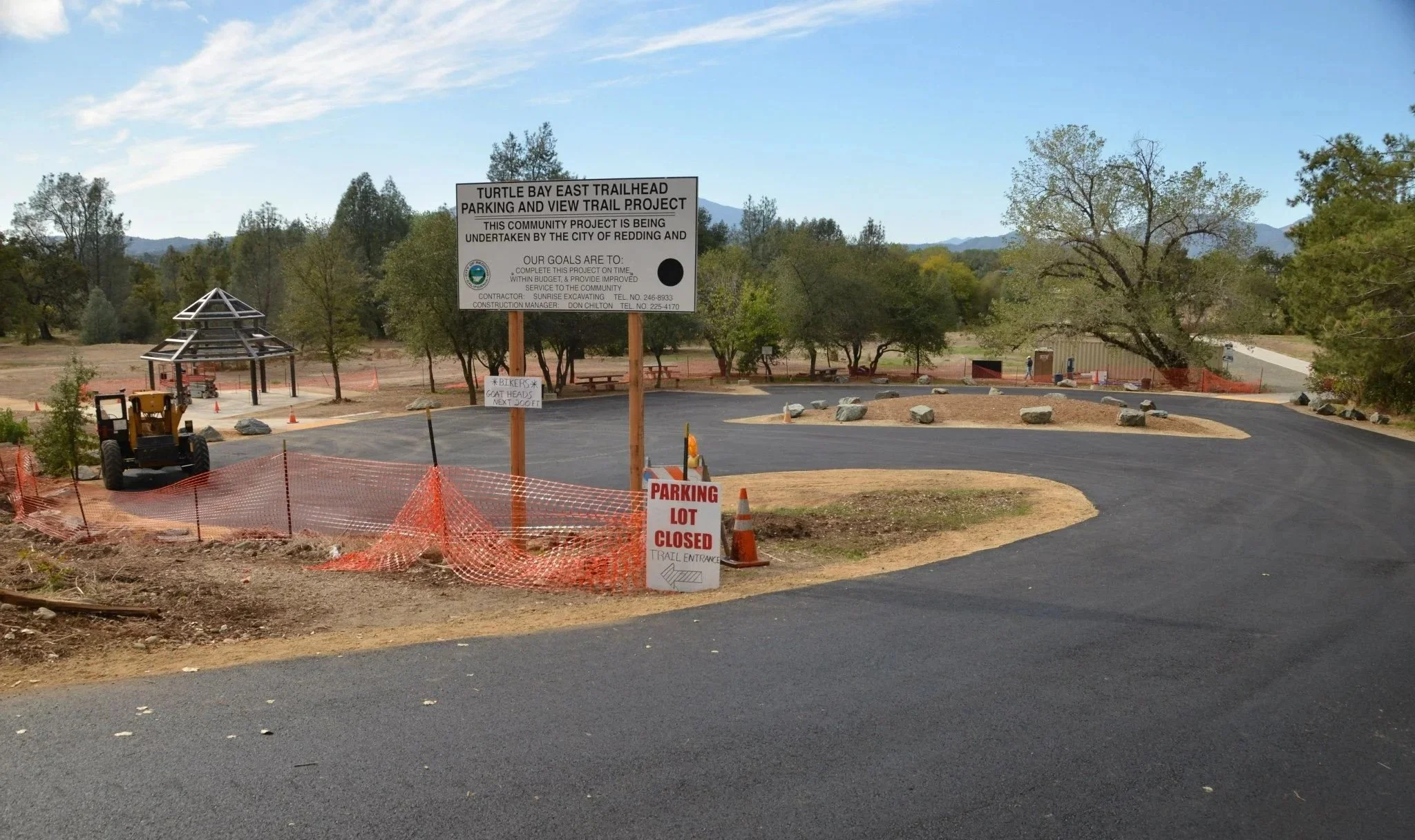 Construction site with a paved road, orange safety fencing, and a sign indicating a trailhead project. There is a small structure being built, a parked construction vehicle, and trees with mountains in the background.