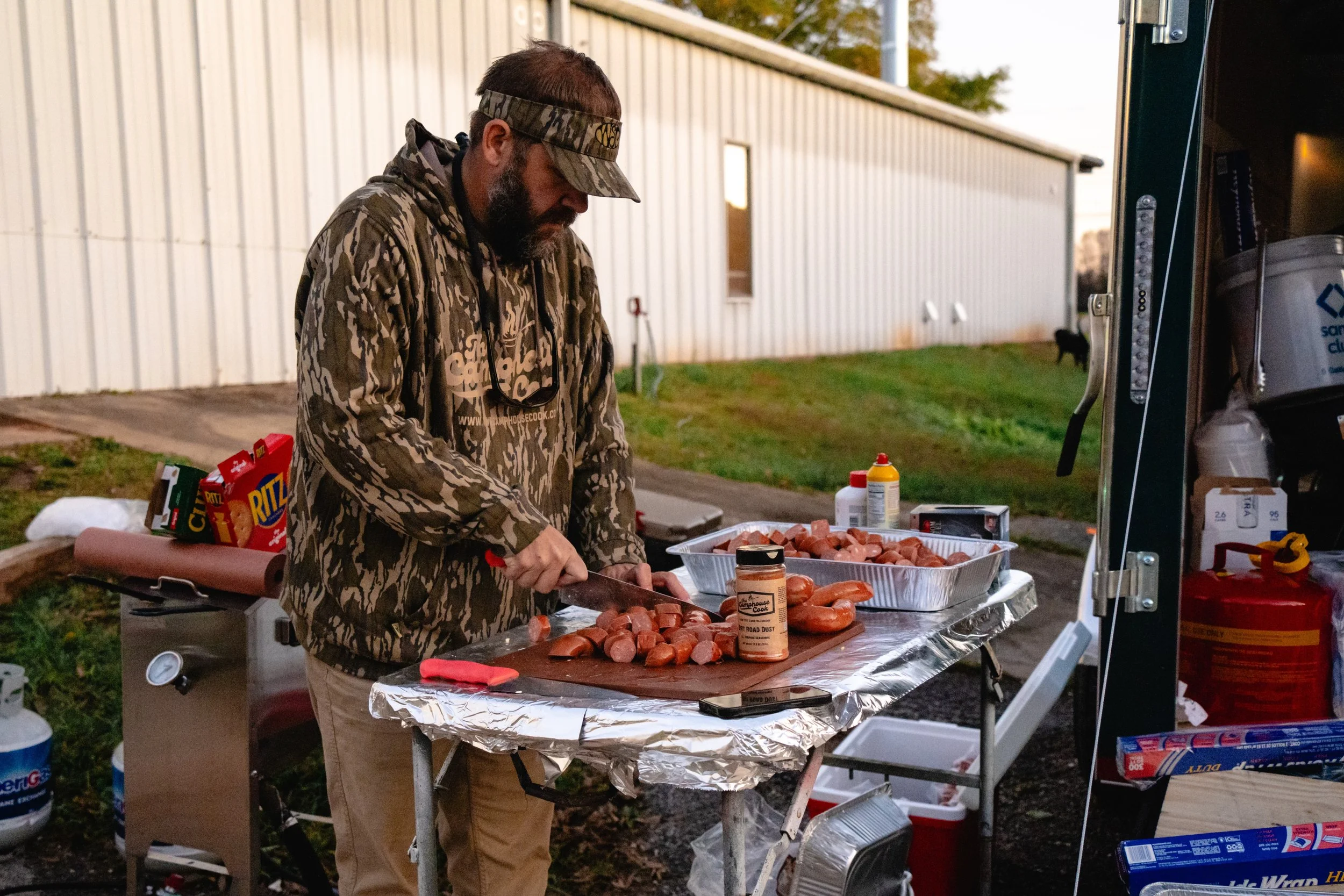 A man with a beard wearing camouflage hoodie and a camo cap preparing sausages at an outdoor grilling station during the evening. There is a container of raw sausages, barbecue spices, and other grilling supplies on the table. The setting appears to 