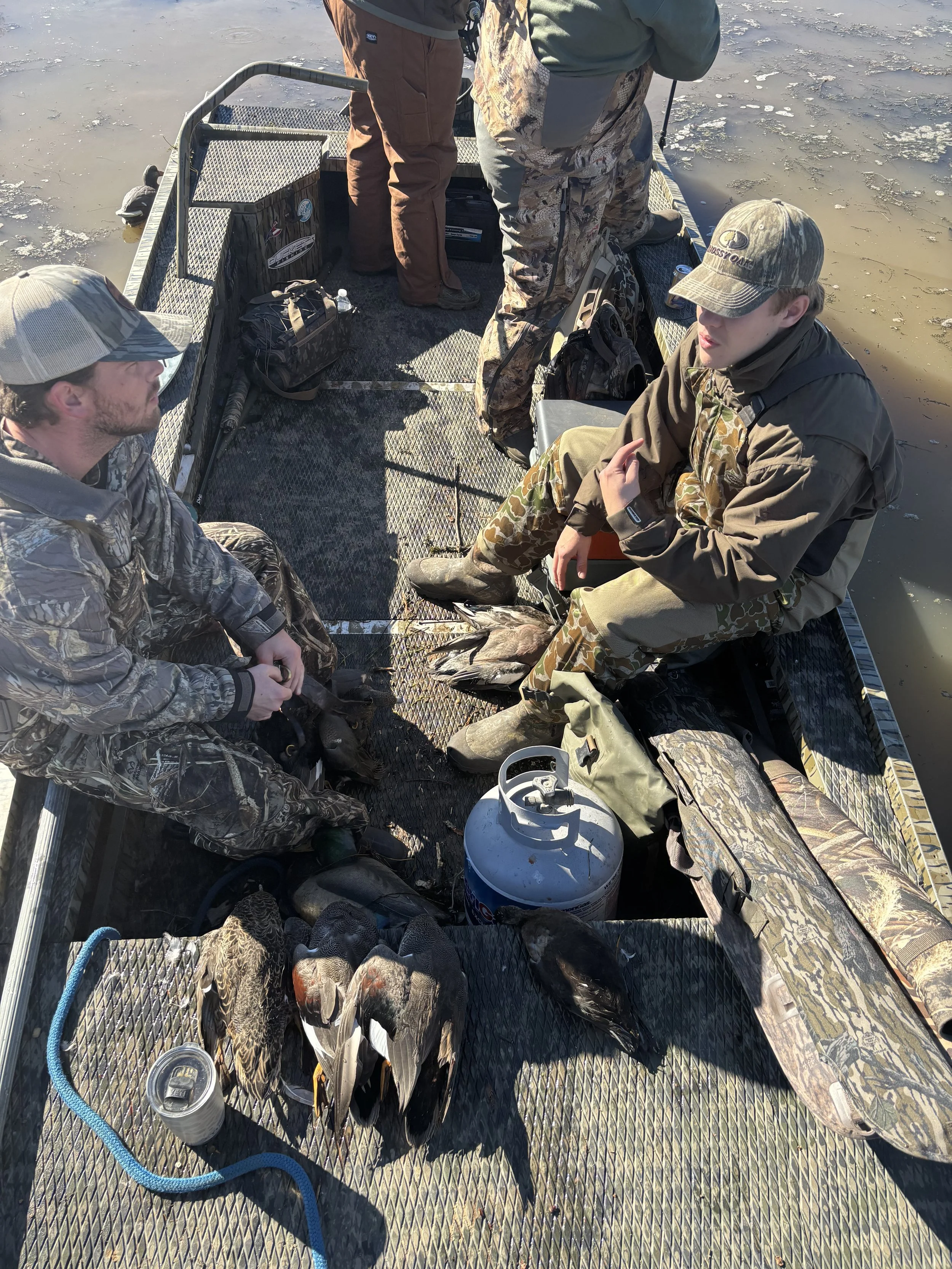 Four people on a boat in a frozen body of water, with fish caught and laid out on the boat's floor, fishing gear, and a cooler visible.