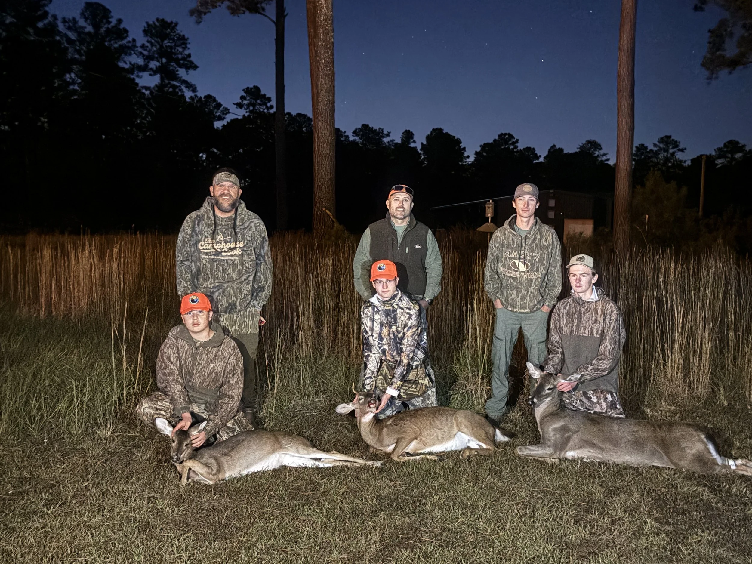 Group of seven people in camouflage outdoor clothing posing outdoors at night, with trees and a starry sky in the background, displaying four deer they hunted with their mounts.