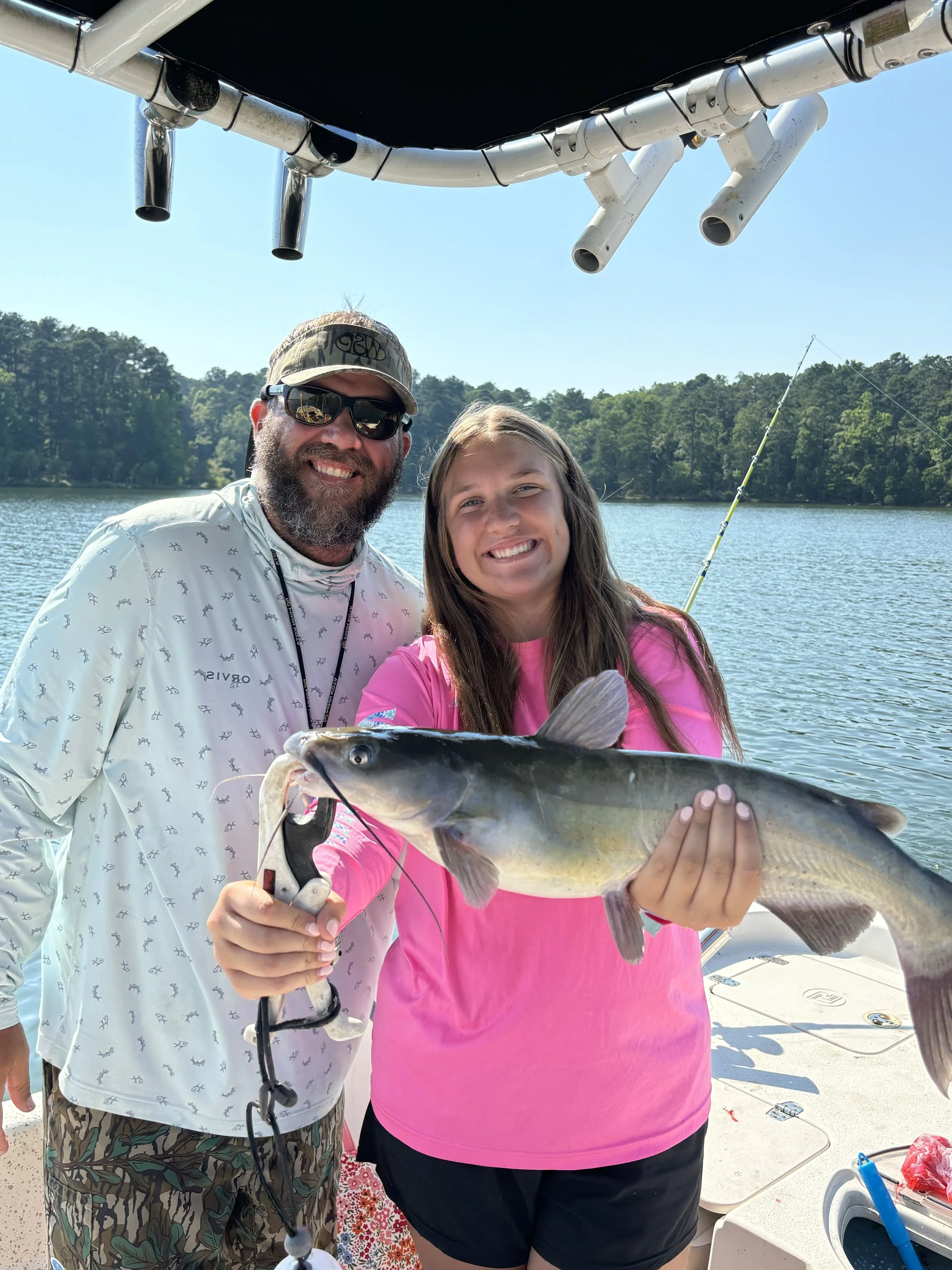 Two people, a man and a woman, are smiling on a boat by a lake. The woman is holding a large fish. The man is wearing sunglasses, a white shirt with small patterns, and camouflage shorts. The woman is wearing a bright pink shirt and black shorts. In the background, there is a fishing rod, water, trees, and a clear blue sky.