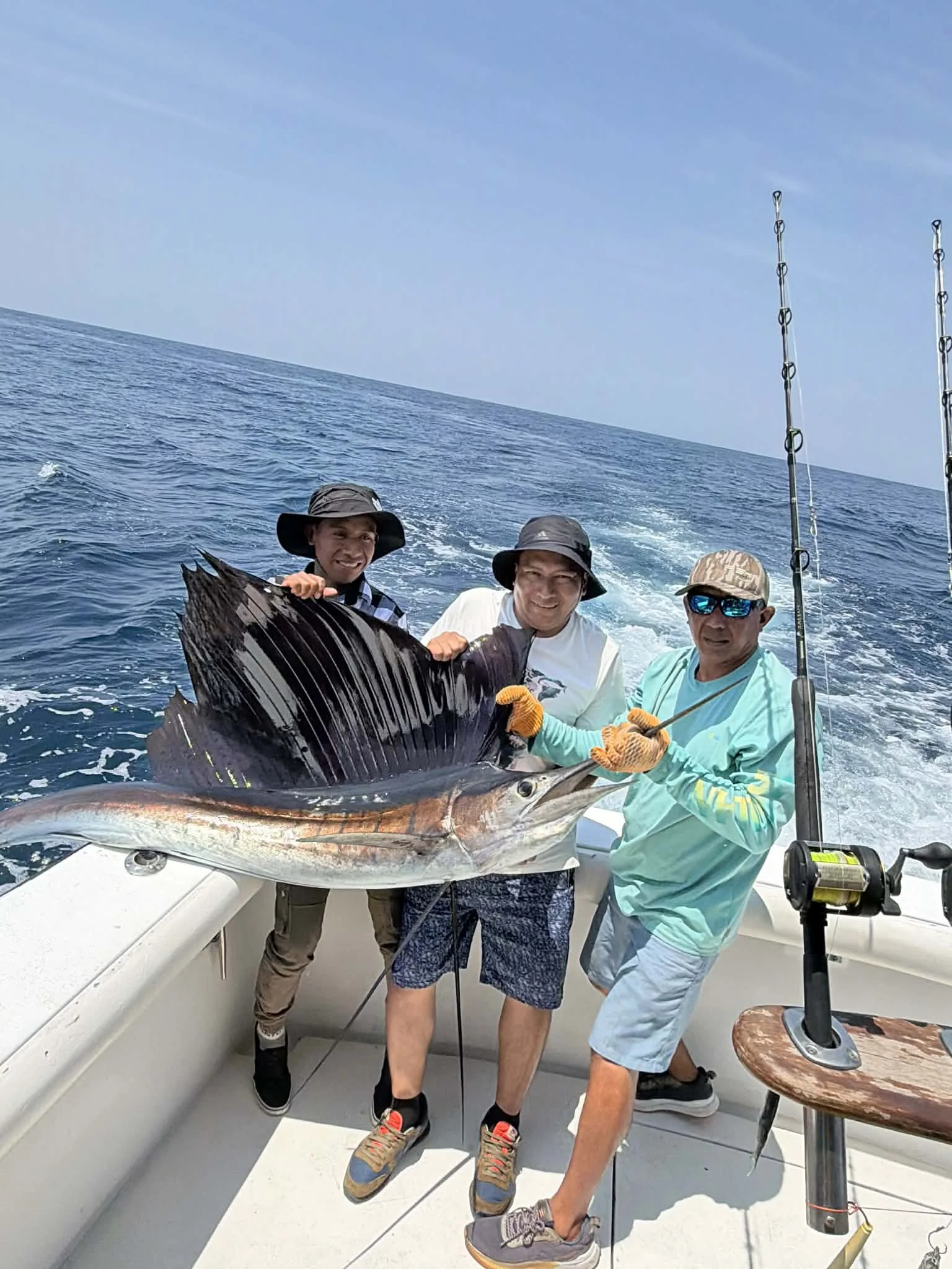 Three men on a boat holding a large fish they caught, with fishing poles visible. The background shows the ocean and a clear blue sky.