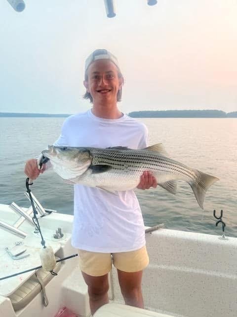 A person on a boat holding a large fish over the water, with a lake and a distant shoreline in the background.