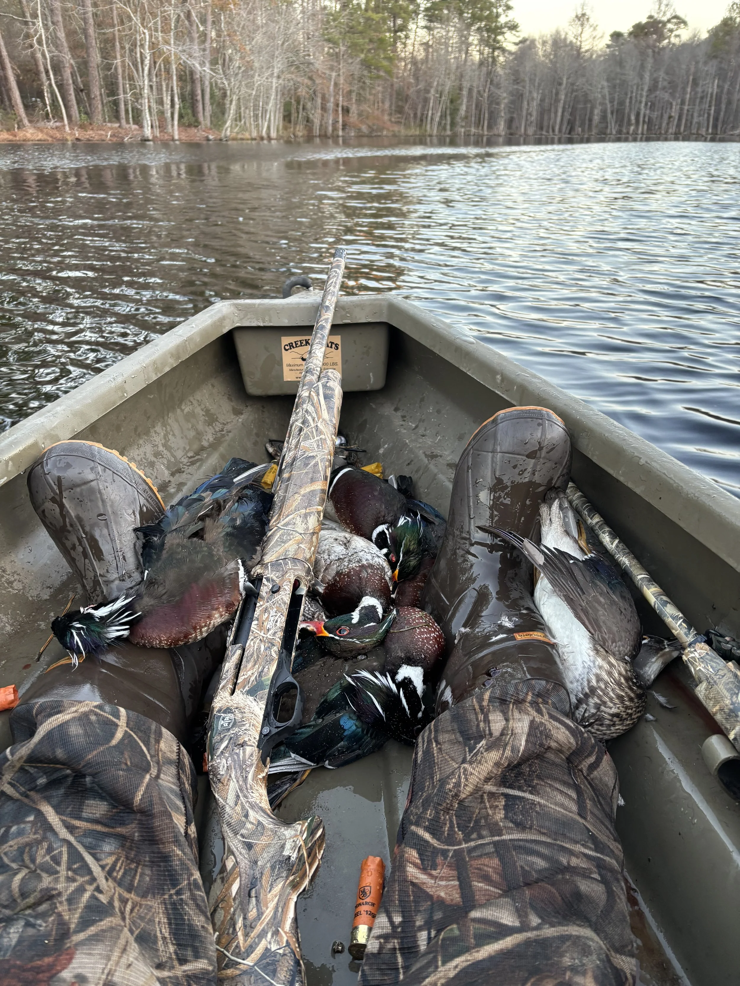 A person sitting in a small boat on a lake, holding multiple ducks and a shotgun, with a wooded shoreline in the background.