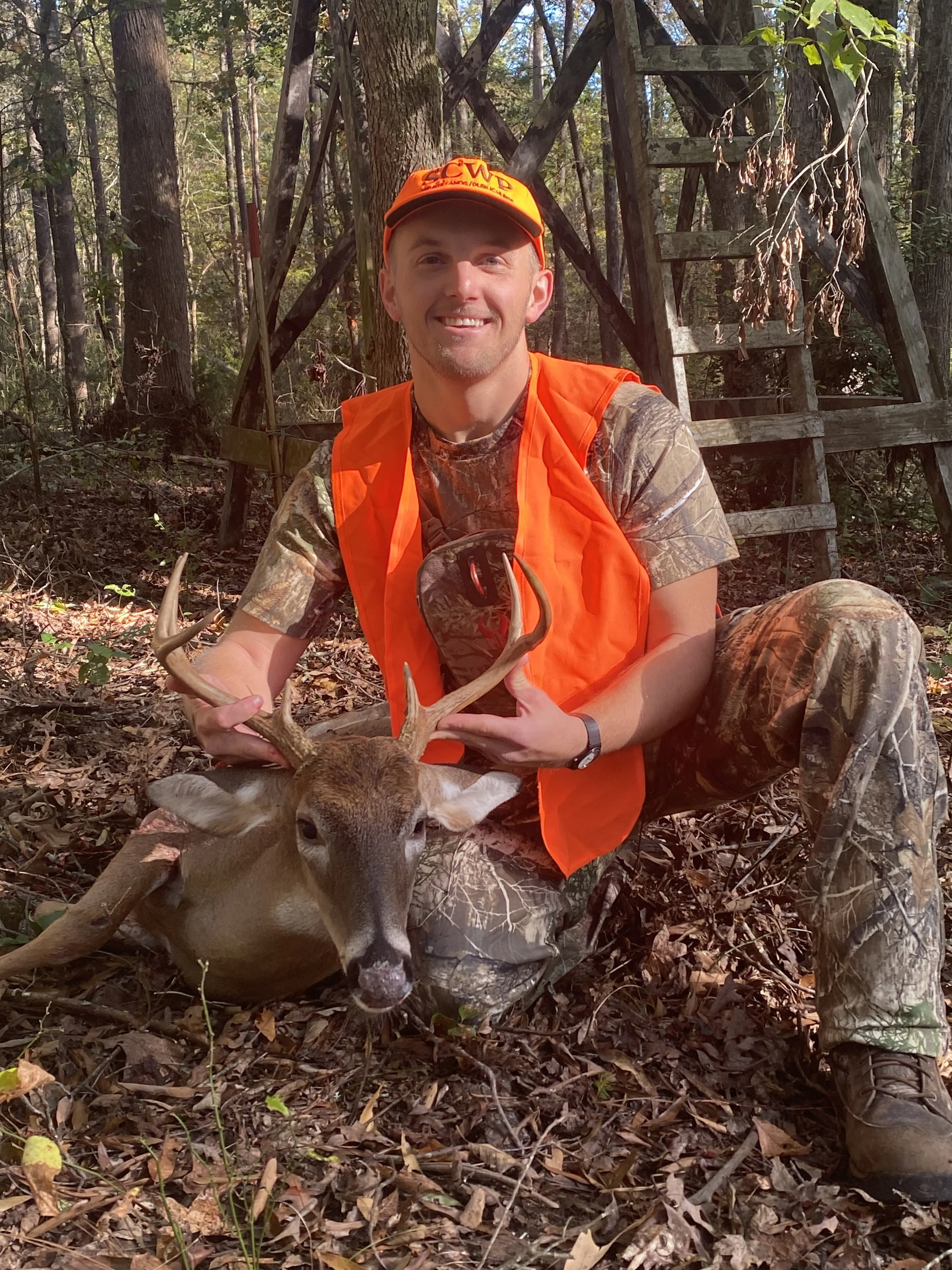 A man wearing camouflage clothing and orange safety gear poses with a deer he has hunted in a wooded area, holding the deer's antlers.