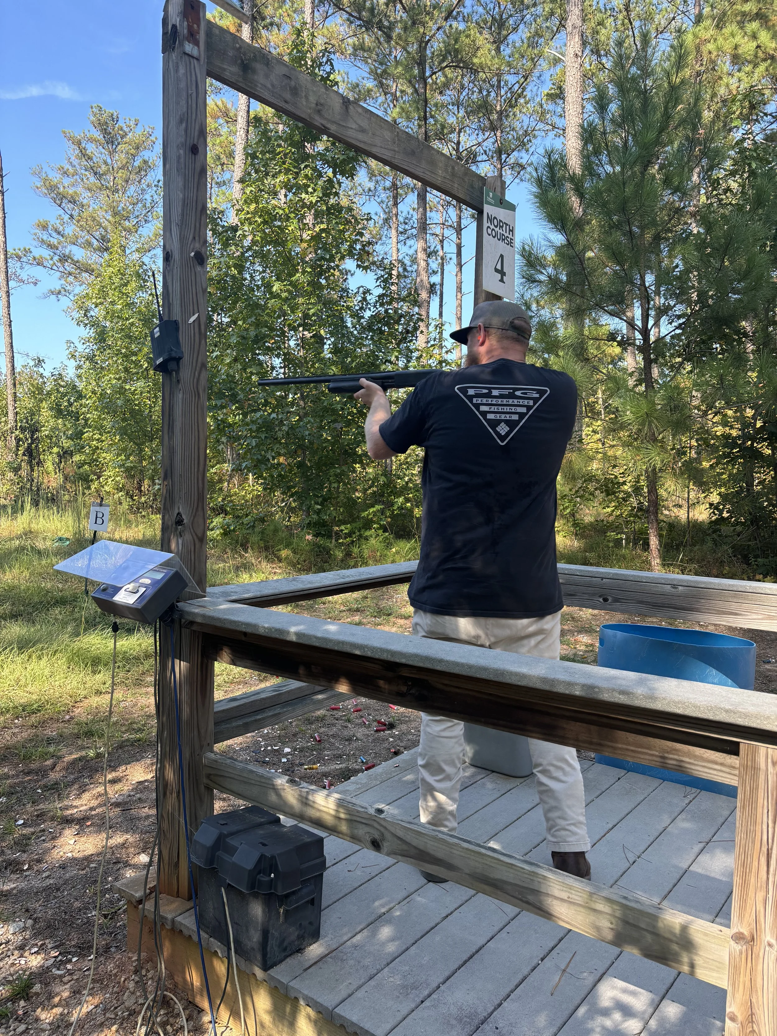 Person shooting clay targets from a shooting platform in a wooded outdoor area, holding a shotgun and wearing a cap and casual clothing.