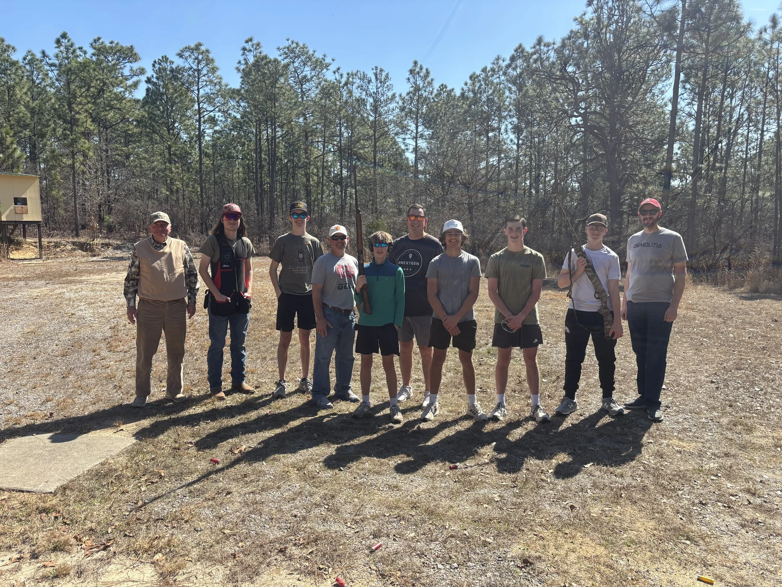 A group of ten people standing outdoors in front of a forested area with tall pine trees, bright blue sky, and a gravel ground. The group includes men, women, and a child, some holding firearms, dressed in casual and outdoor clothing, and smiling at the camera.