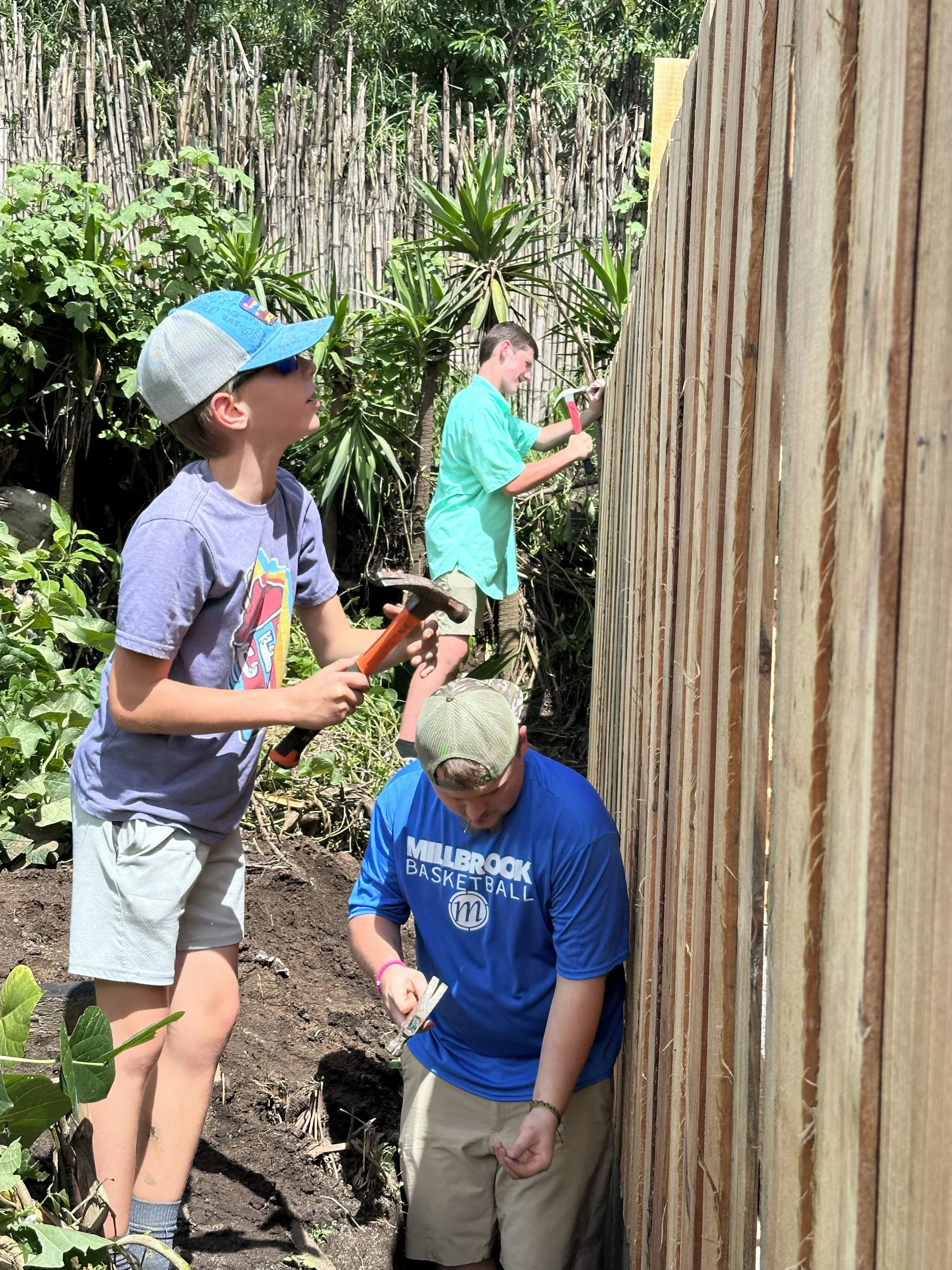 Two children and an adult working together on a gardening project outdoors. The children are holding tools, and the adult is hammering something into a wooden fence. They are surrounded by green plants and a natural setting with a wooden fence in the background.