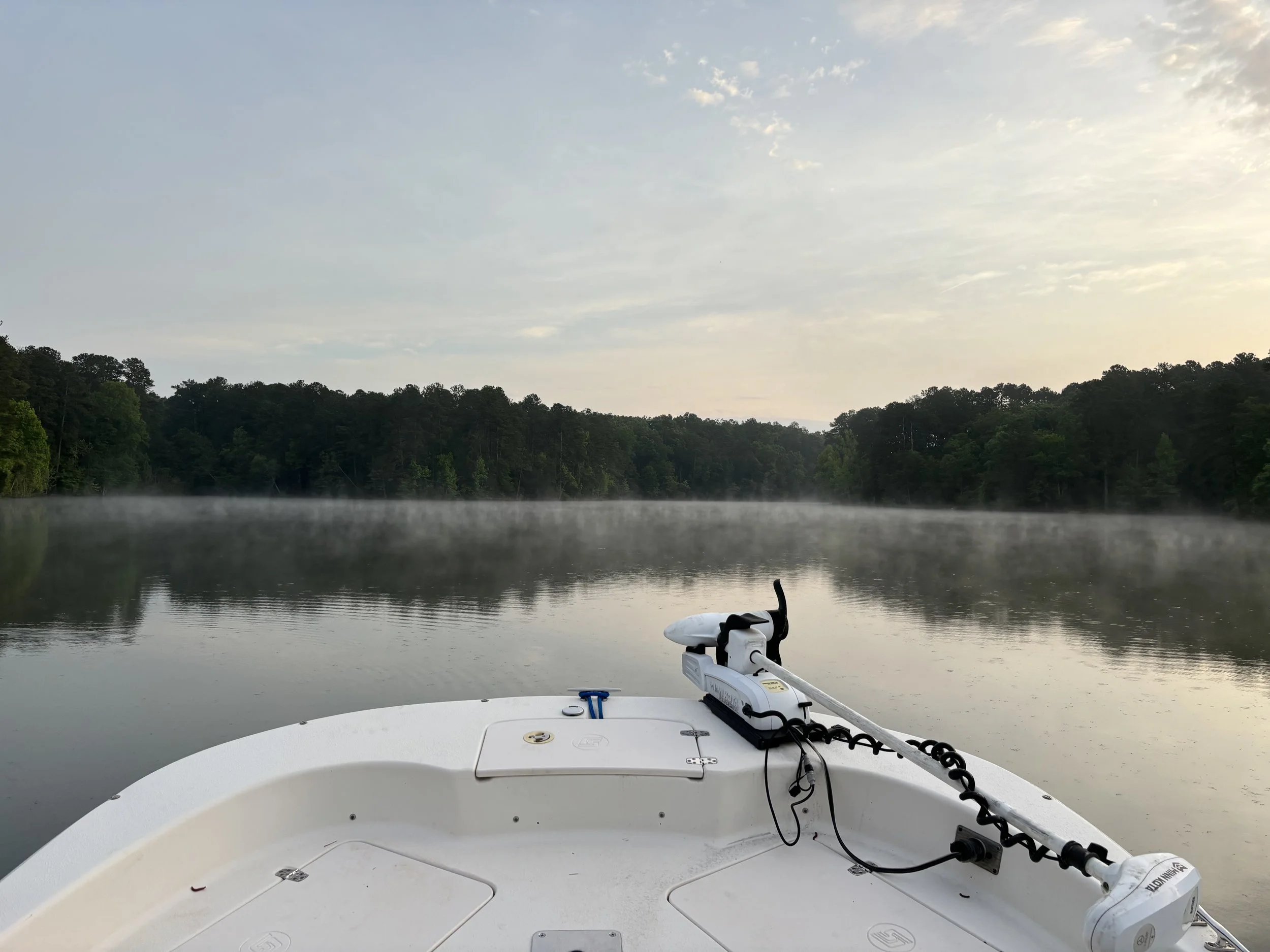 View from a boat on a calm river with fog rising from the water and a forested shoreline in the distance, cloudy sky, and motorboat parts visible in the foreground.