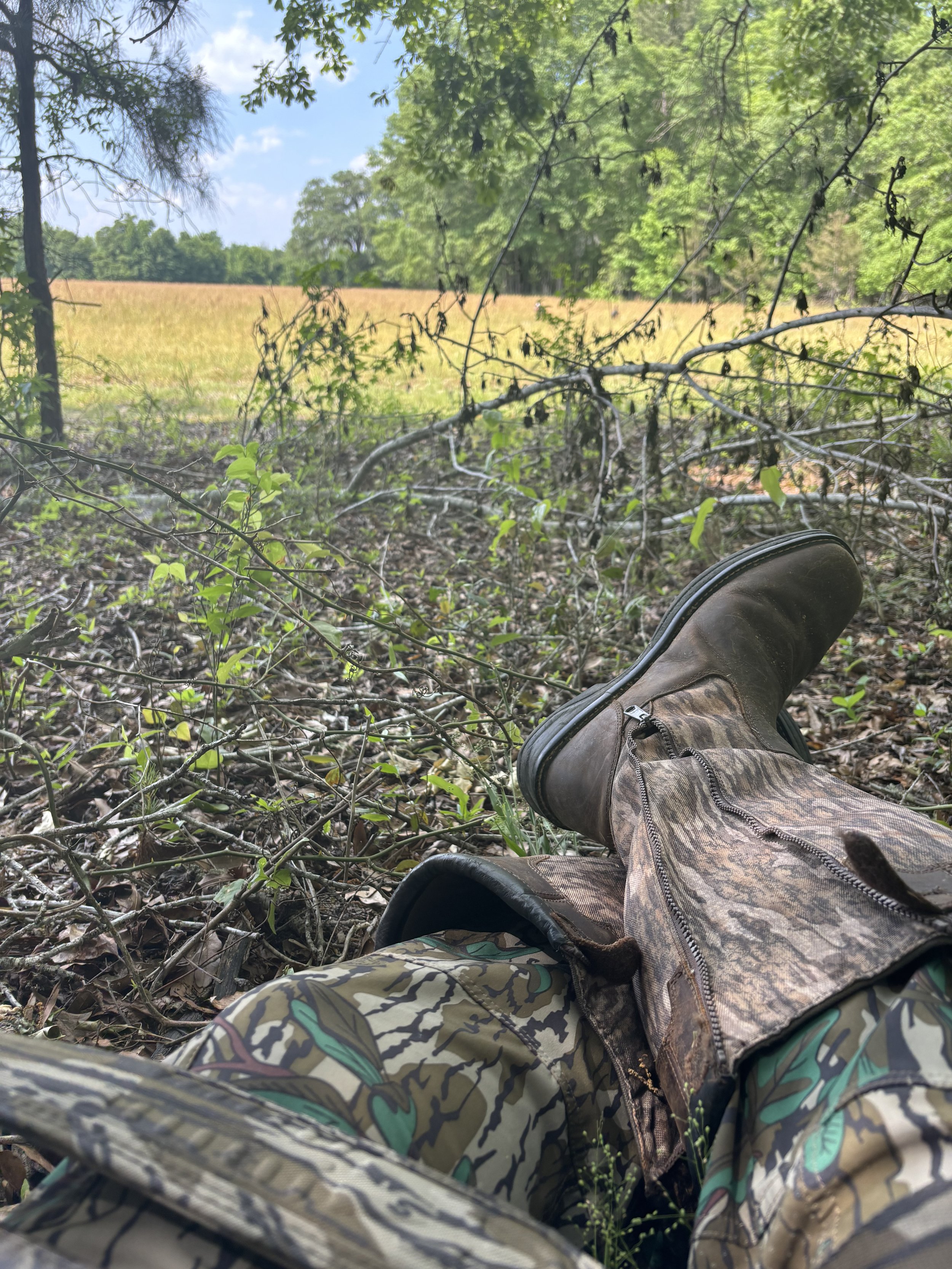 View of a person resting outdoors, with their legs extended on the ground, wearing camouflage pants and brown boots. The background shows a field with green trees and a clear blue sky.