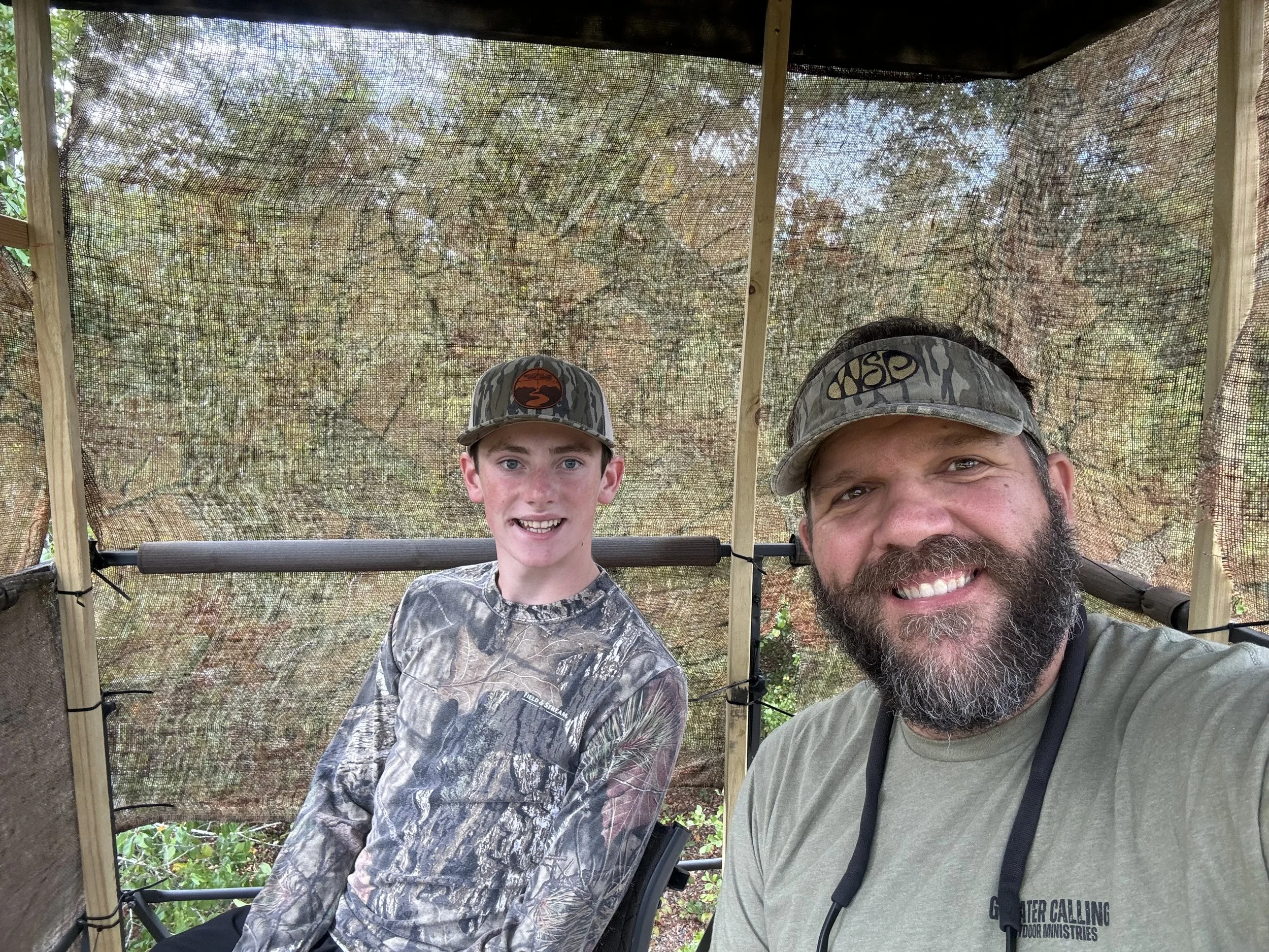 A man and a boy are smiling for a selfie inside a shaded outdoor shelter with a mesh roof. Both are wearing camouflage hats, and the man is wearing a light-colored shirt with black straps, possibly from a camera or equipment. There is greenery outsid
