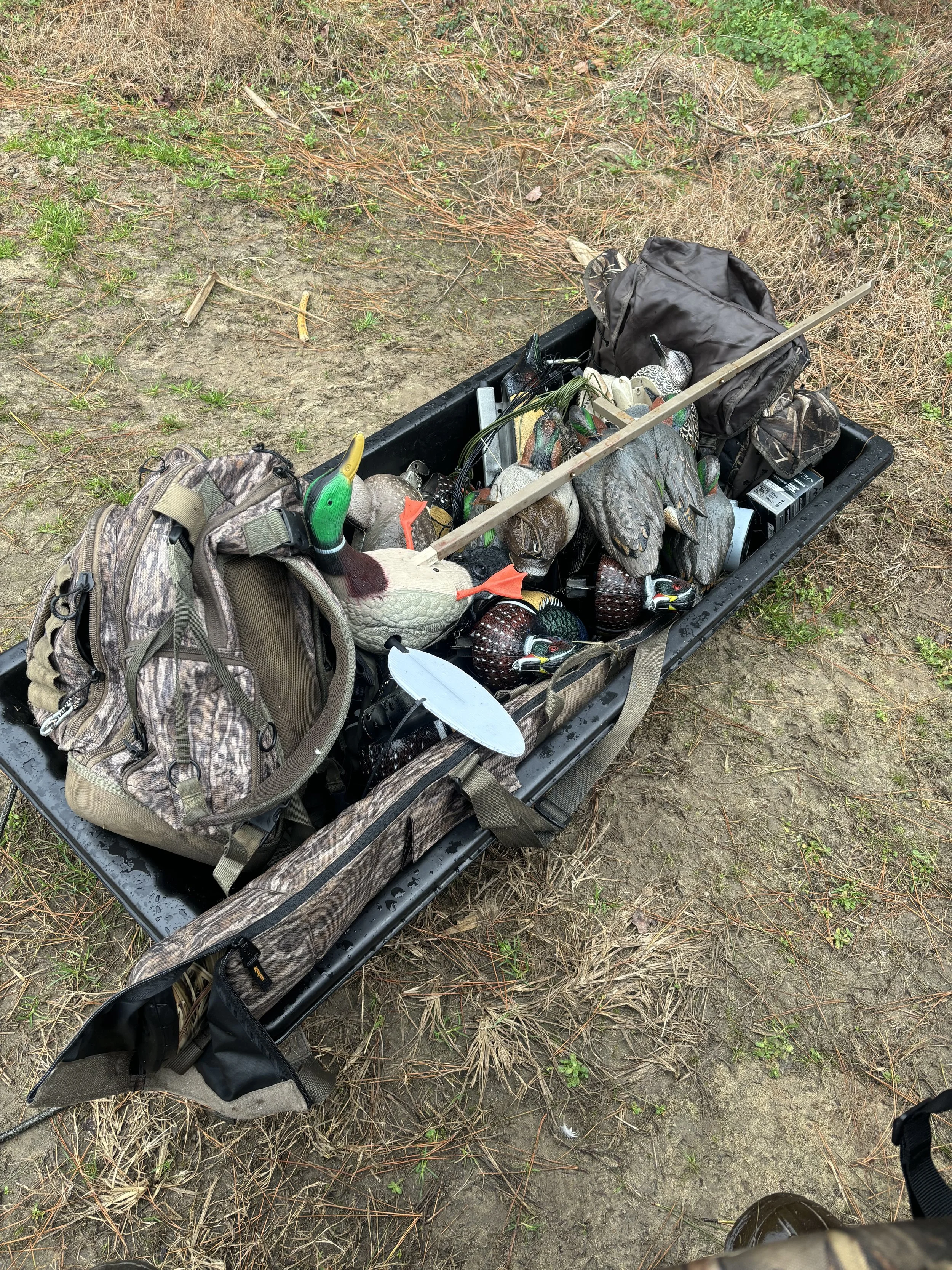 A black plastic kayak filled with hunting gear, including decoys, a camouflage backpack, and other supplies, resting on dirt ground with some grass and dry foliage around.