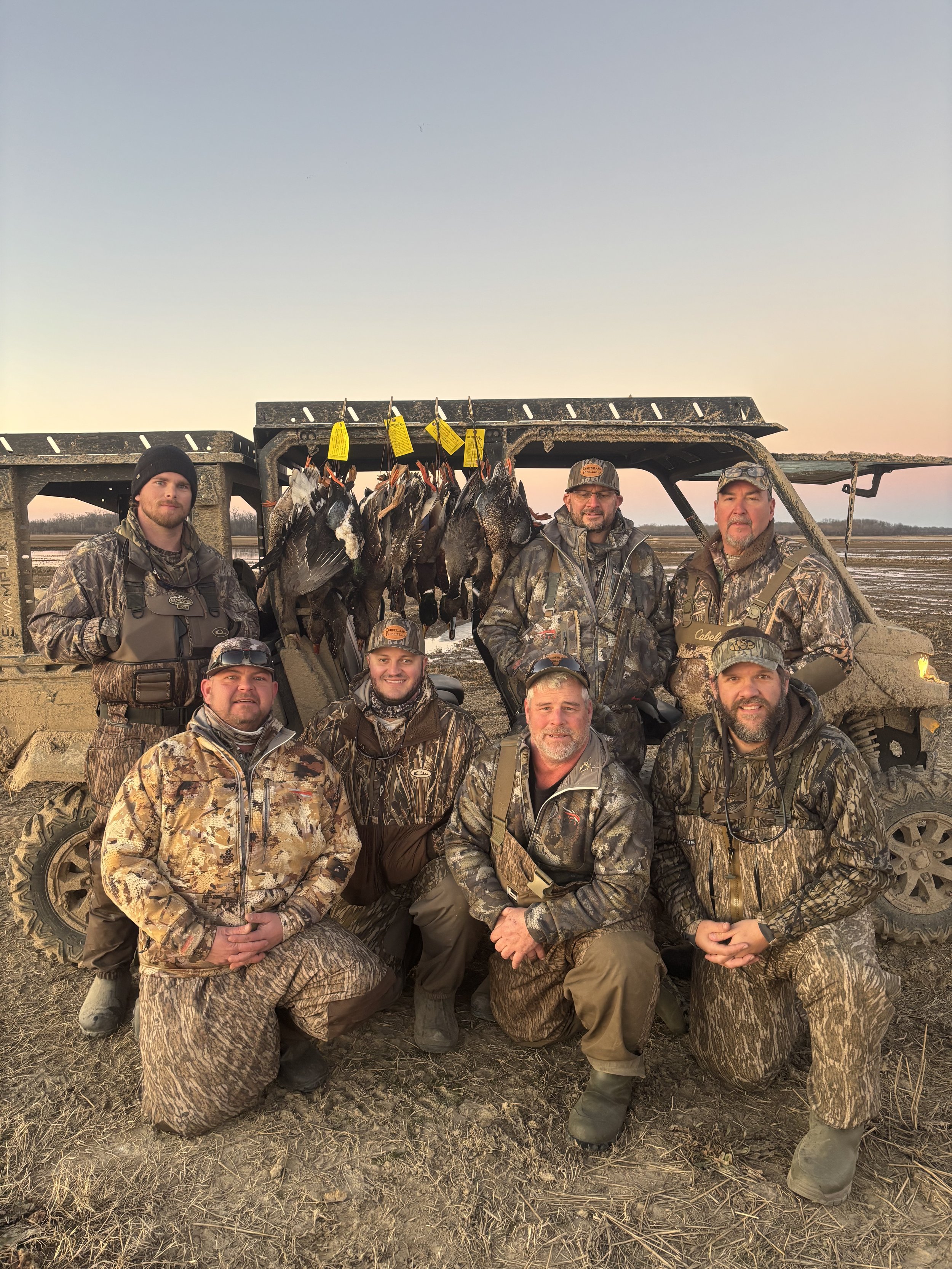A group of five men in camouflage clothing and gear, posing in front of a muddy field and a vehicle, with a rack of dead ducks hanging nearby, during sunset.