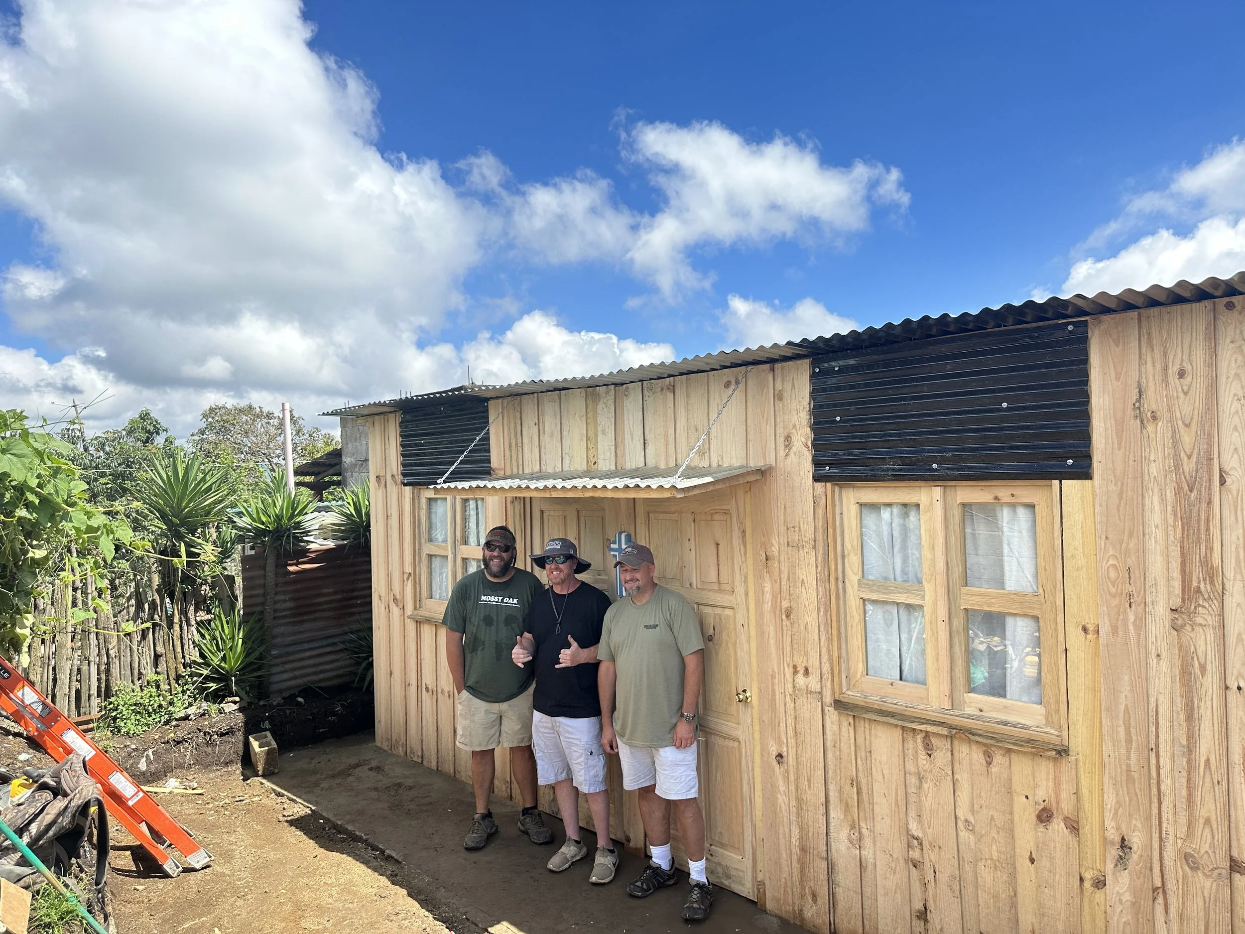 Four men are standing in front of a new wooden building with a corrugated metal roof, under a partly cloudy sky. They are smiling and posing for the photo.