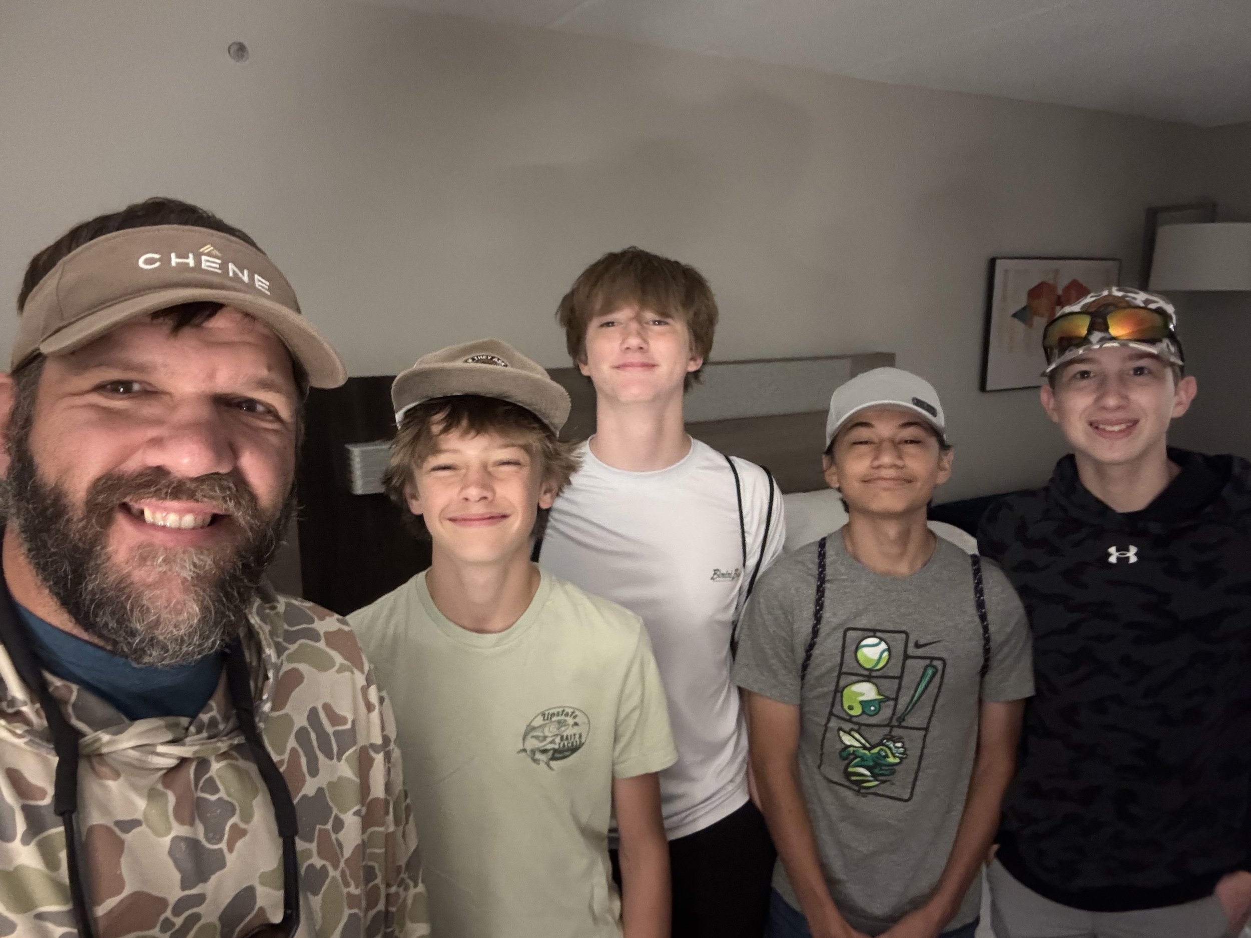 Group of six people smiling and posing indoors, with one man on the left wearing a tan baseball cap and camouflage jacket, and five children, some wearing hats and sunglasses, standing in front of a plain wall with a framed picture.