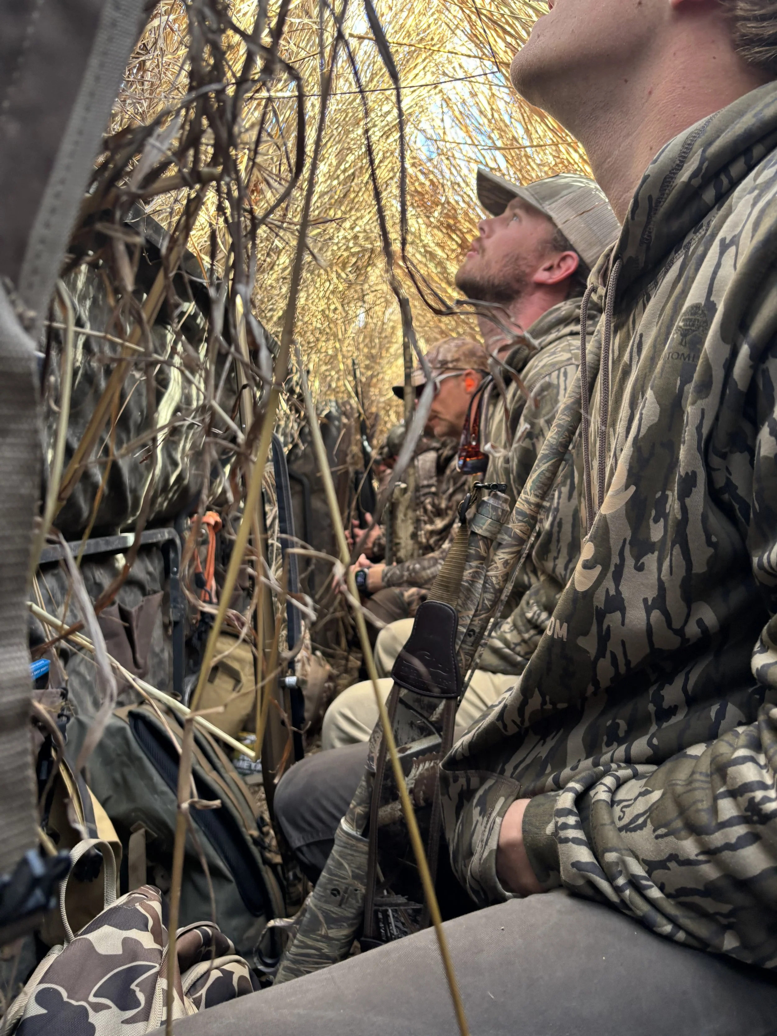 People sitting closely together with camouflage clothing and gear, possibly during a hunting or outdoor activity, with a view of the sky and trees above.