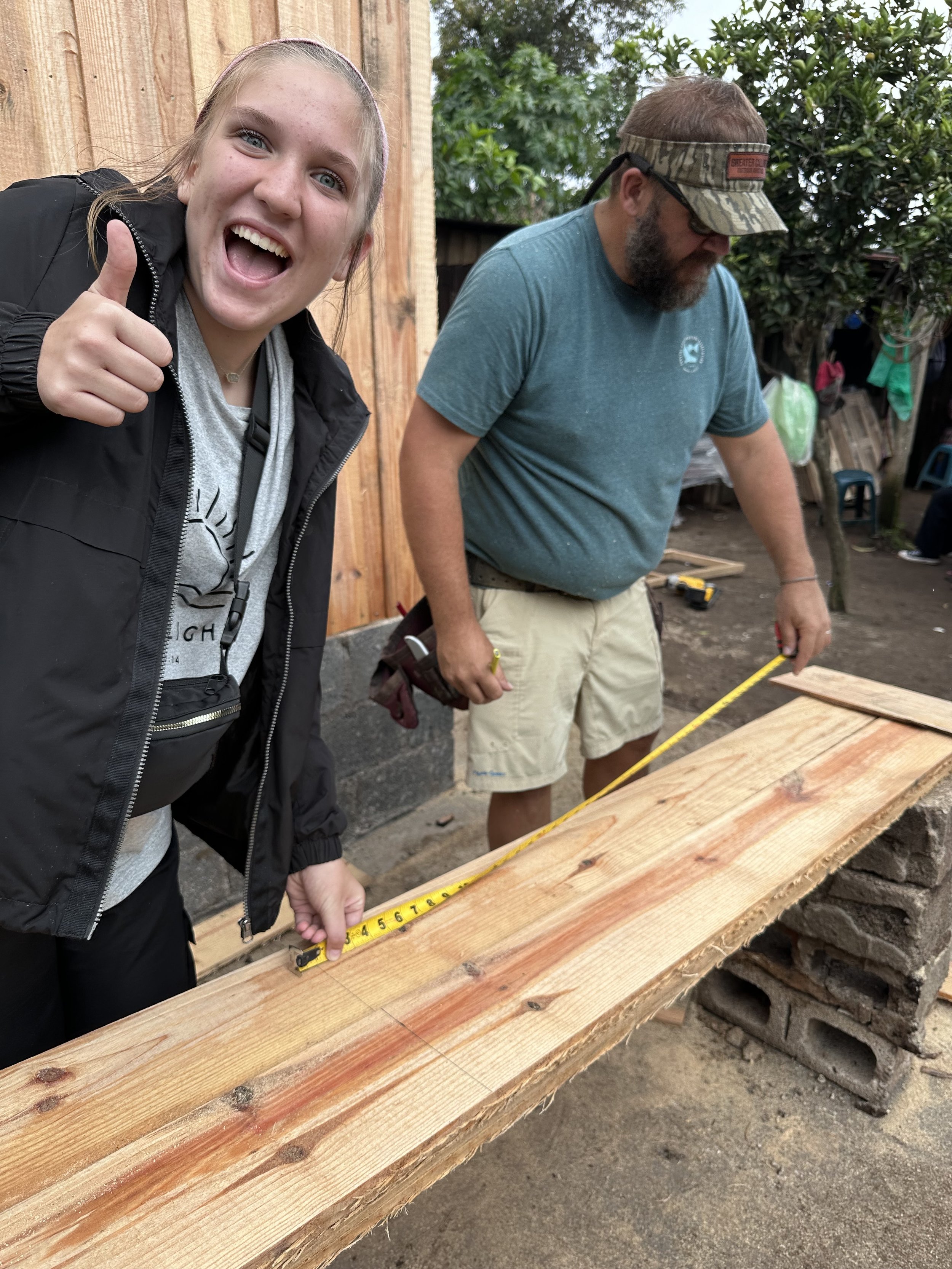 A young woman smiling and giving a thumbs up while holding a measuring tape at a woodworking site, with a man working on a wooden plank nearby. The background features a wooden fence, trees, and various tools.