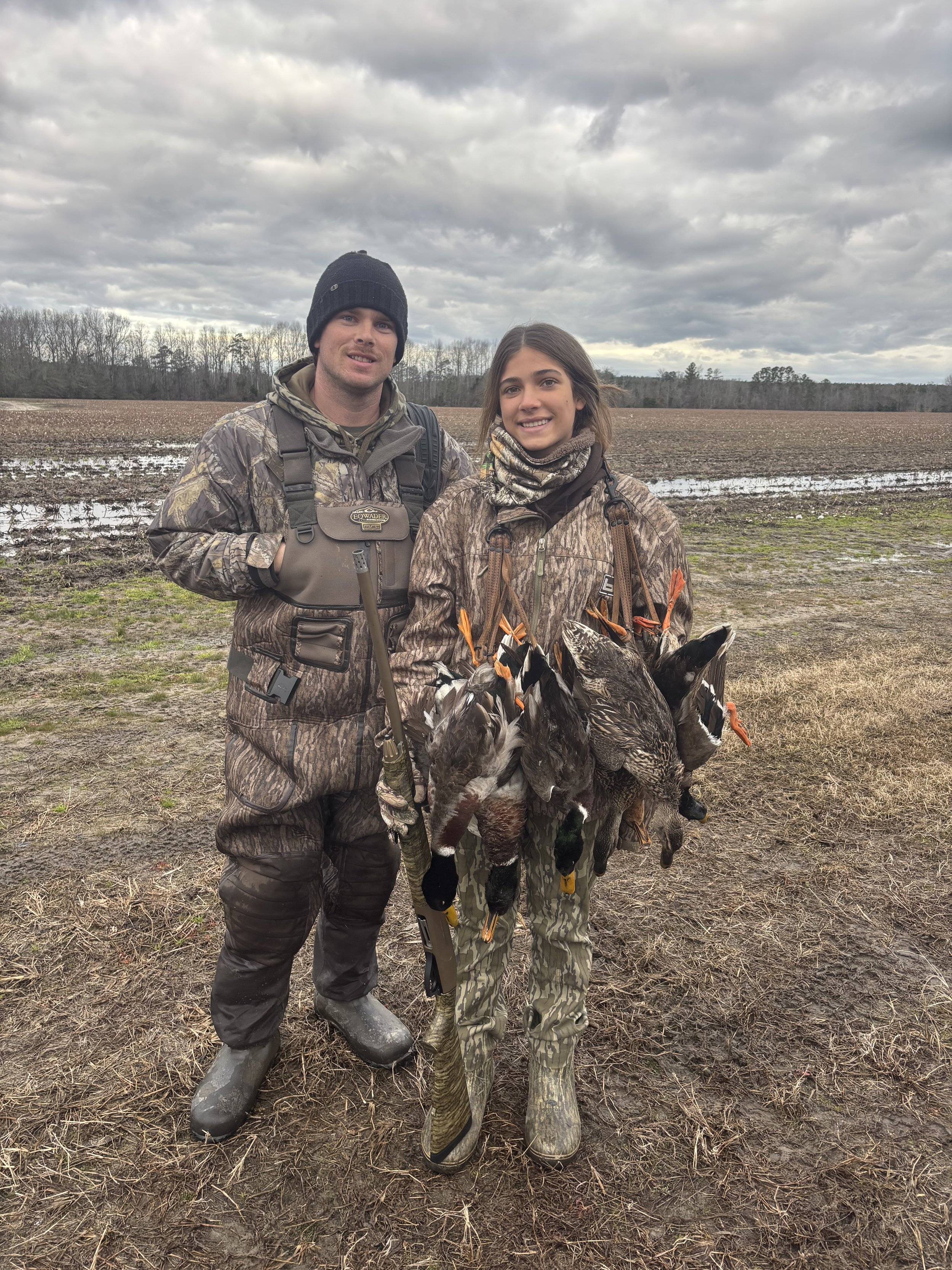 A man and a woman standing outdoors in a muddy field, holding multiple ducks with orange tags attached to their legs. The man is wearing camouflage clothing, a black beanie, and rubber boots. The woman is dressed in camouflage clothing and a neck gaiter. The overcast sky is cloudy, and a line of trees can be seen in the background.