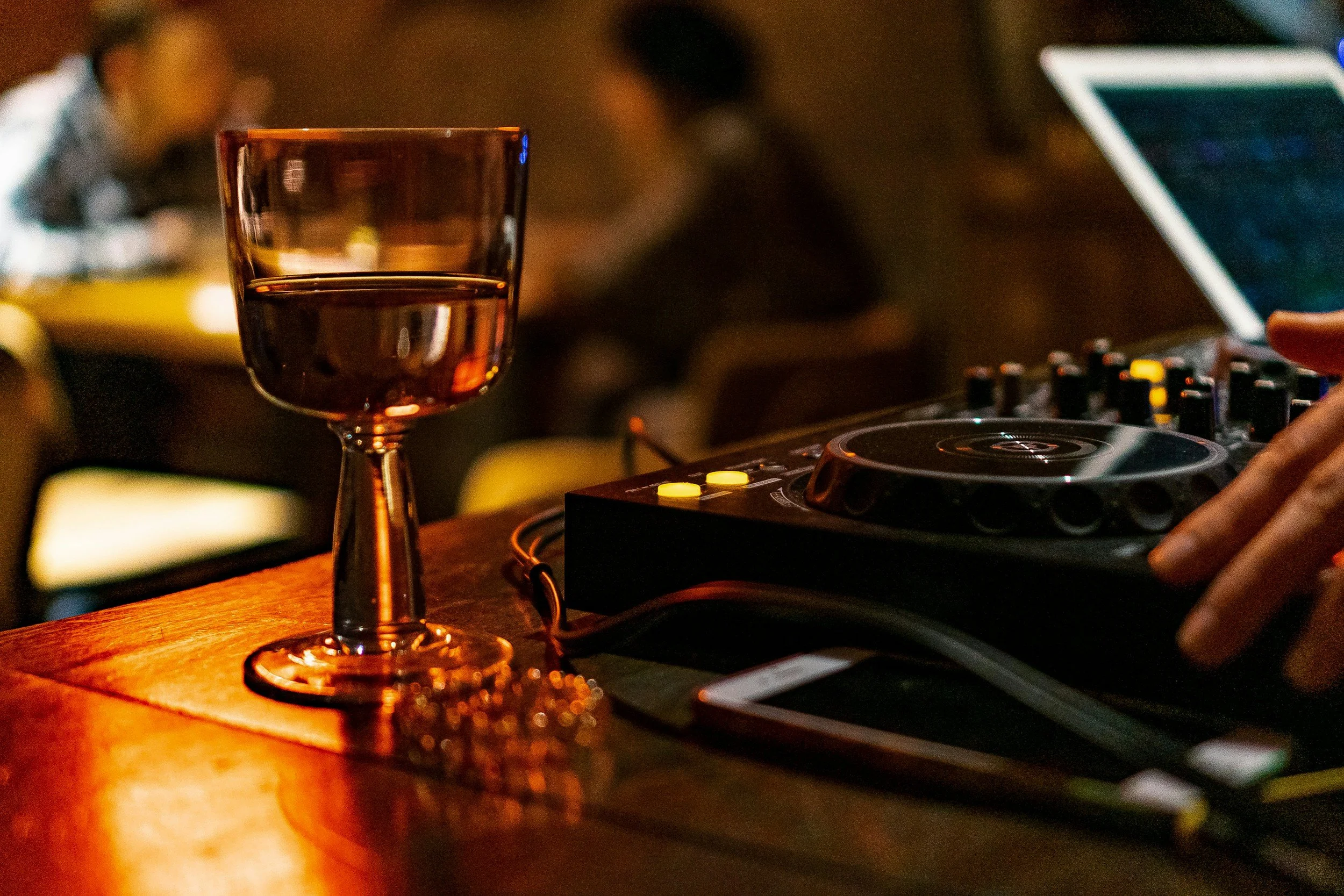 A glass of red wine on a wooden table with a DJ mixer and a laptop in the background, suggesting a party or music event.