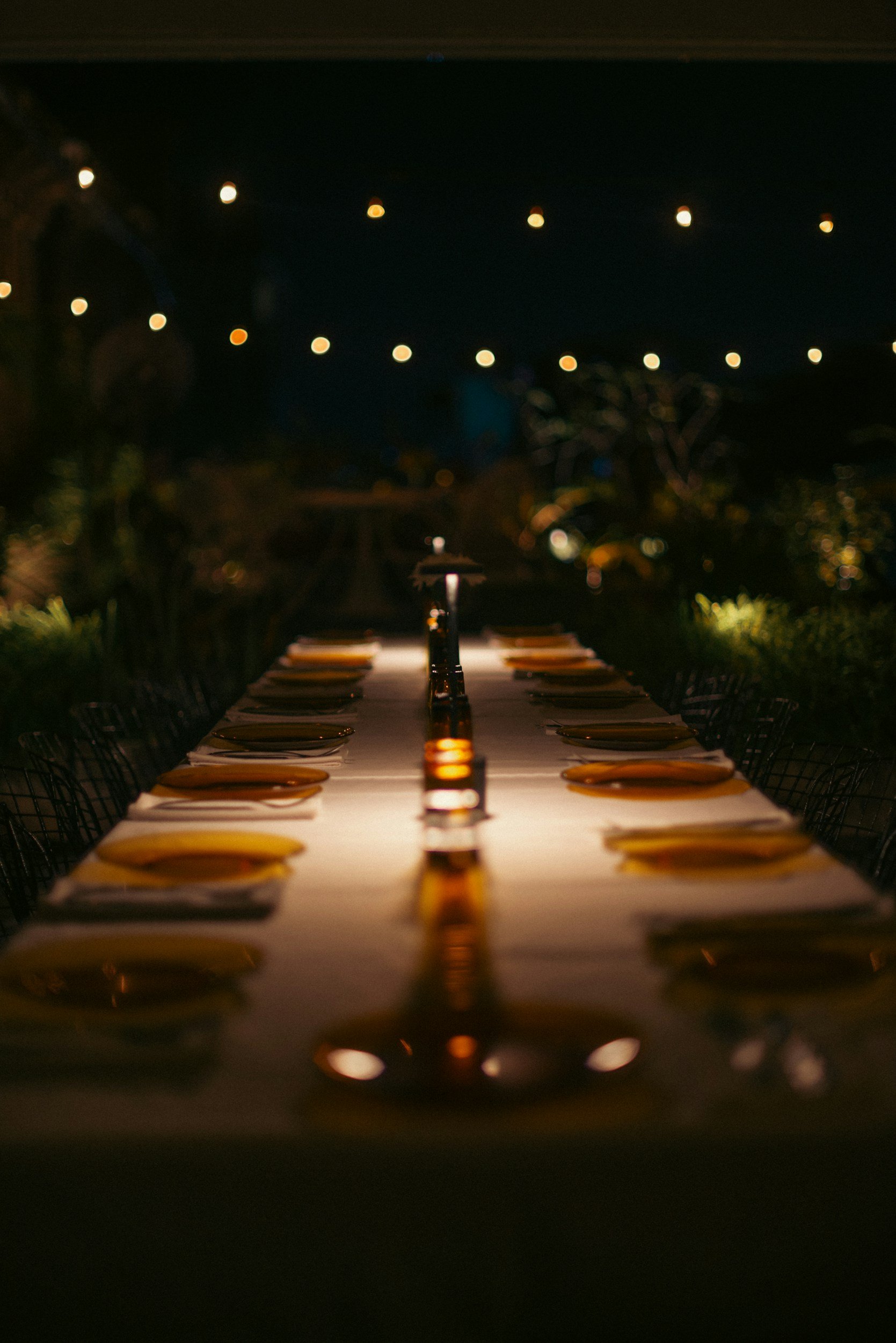 A long dinner table set with plates and utensils, illuminated by string lights overhead, in an outdoor night setting.