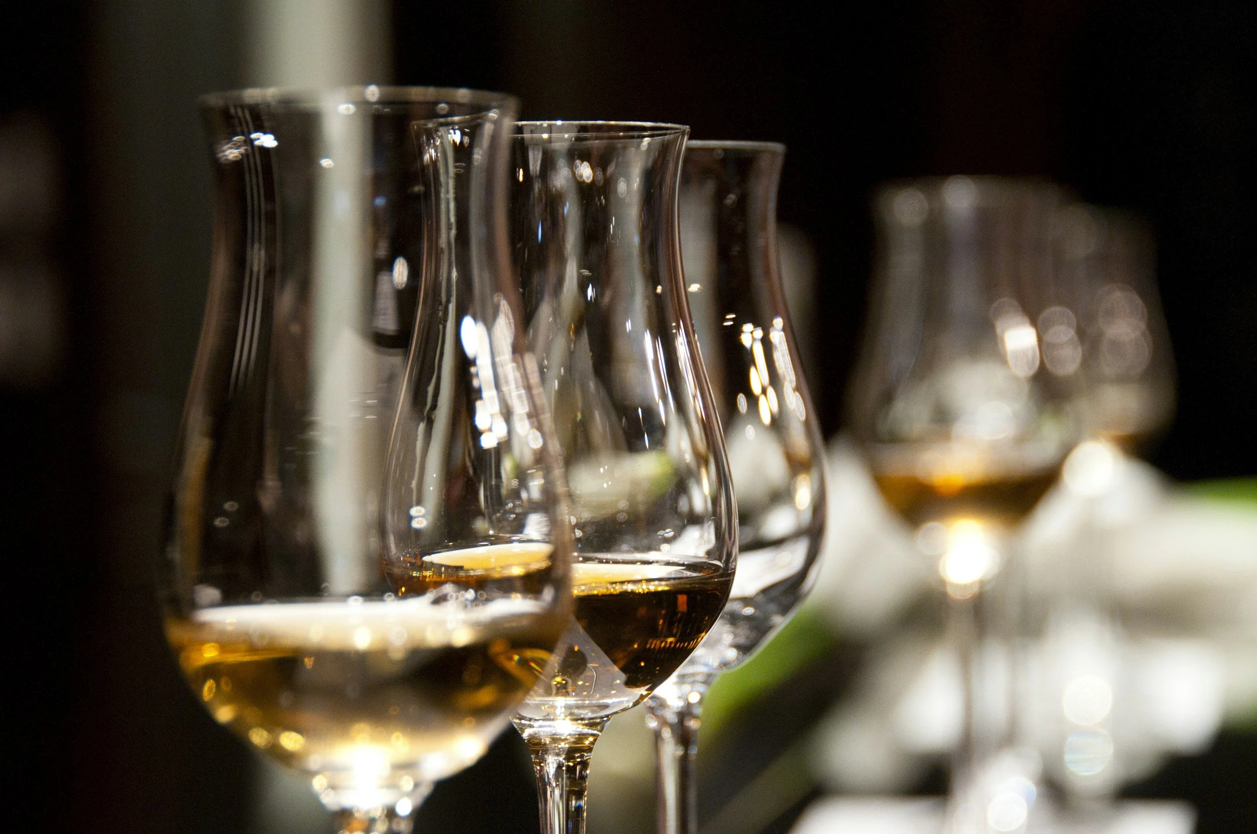 Multiple glasses of amber-colored liquid, likely whiskey, arranged in a line on a dark background with soft lighting and reflections.