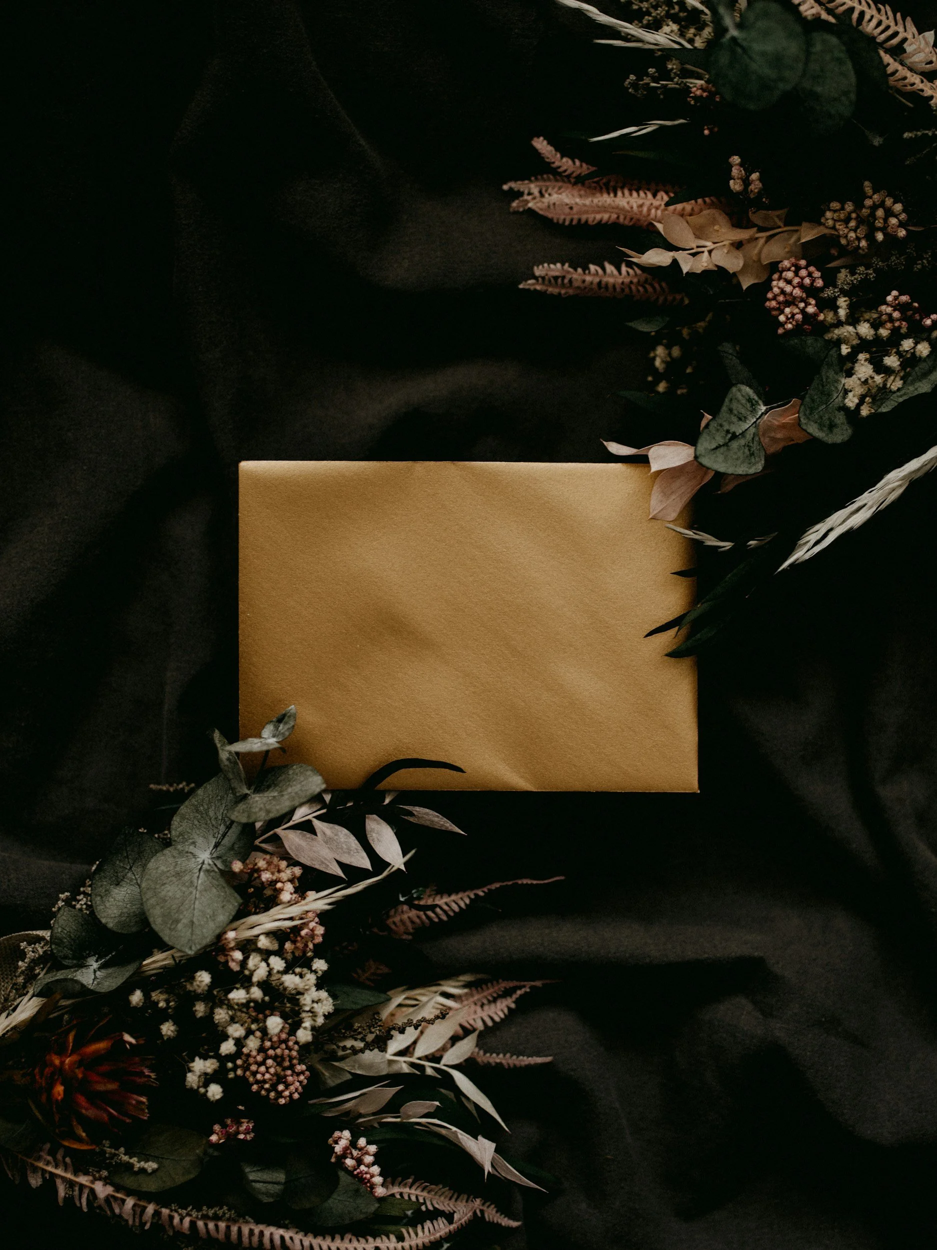 A black cloth background with a gold-colored rectangular gift box surrounded by a bouquet of dried flowers and foliage, including ferns, eucalyptus, and other assorted dried botanical elements.