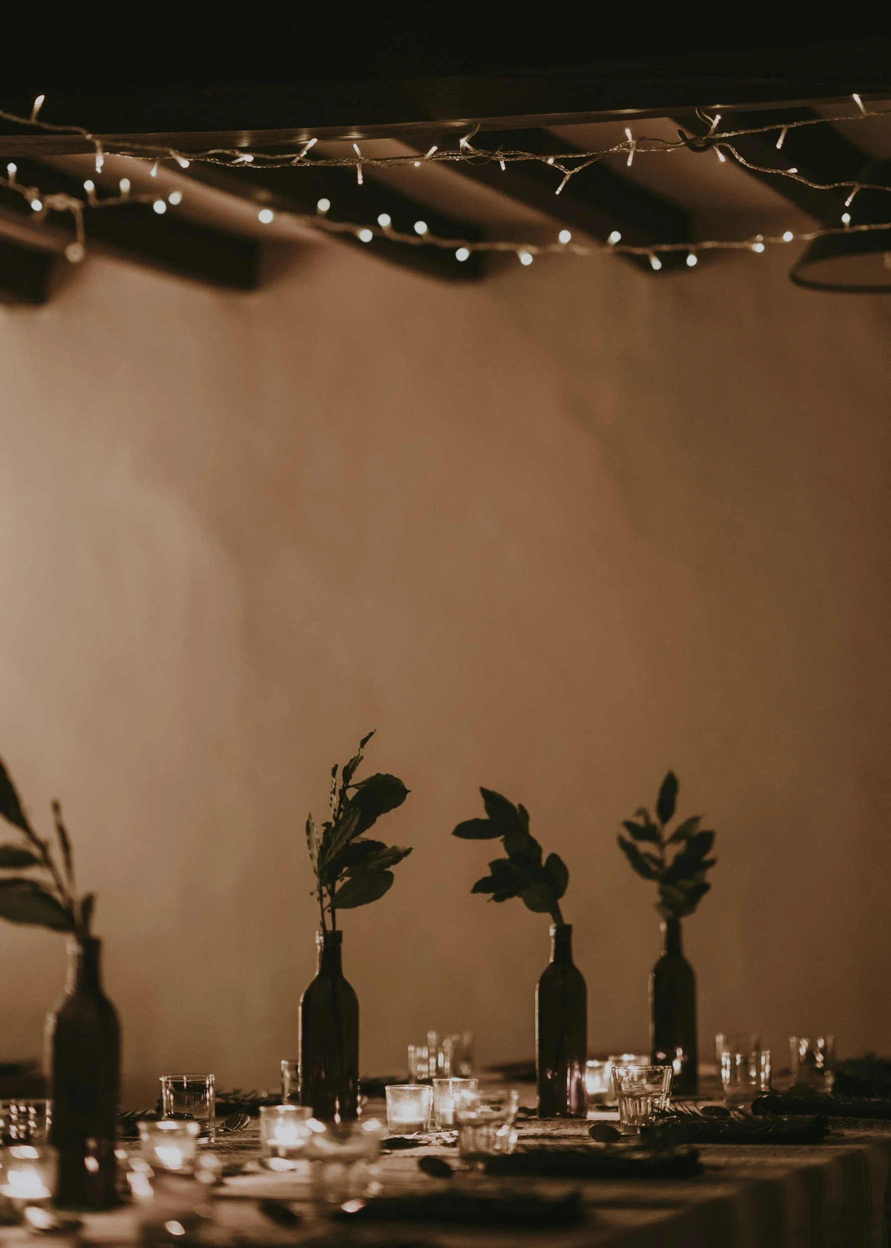 Table decorated with candles, glassware, and tall bottles with green plants or flowers in dark bottles, illuminated by small candles and fairy lights above, set for a cozy evening gathering.