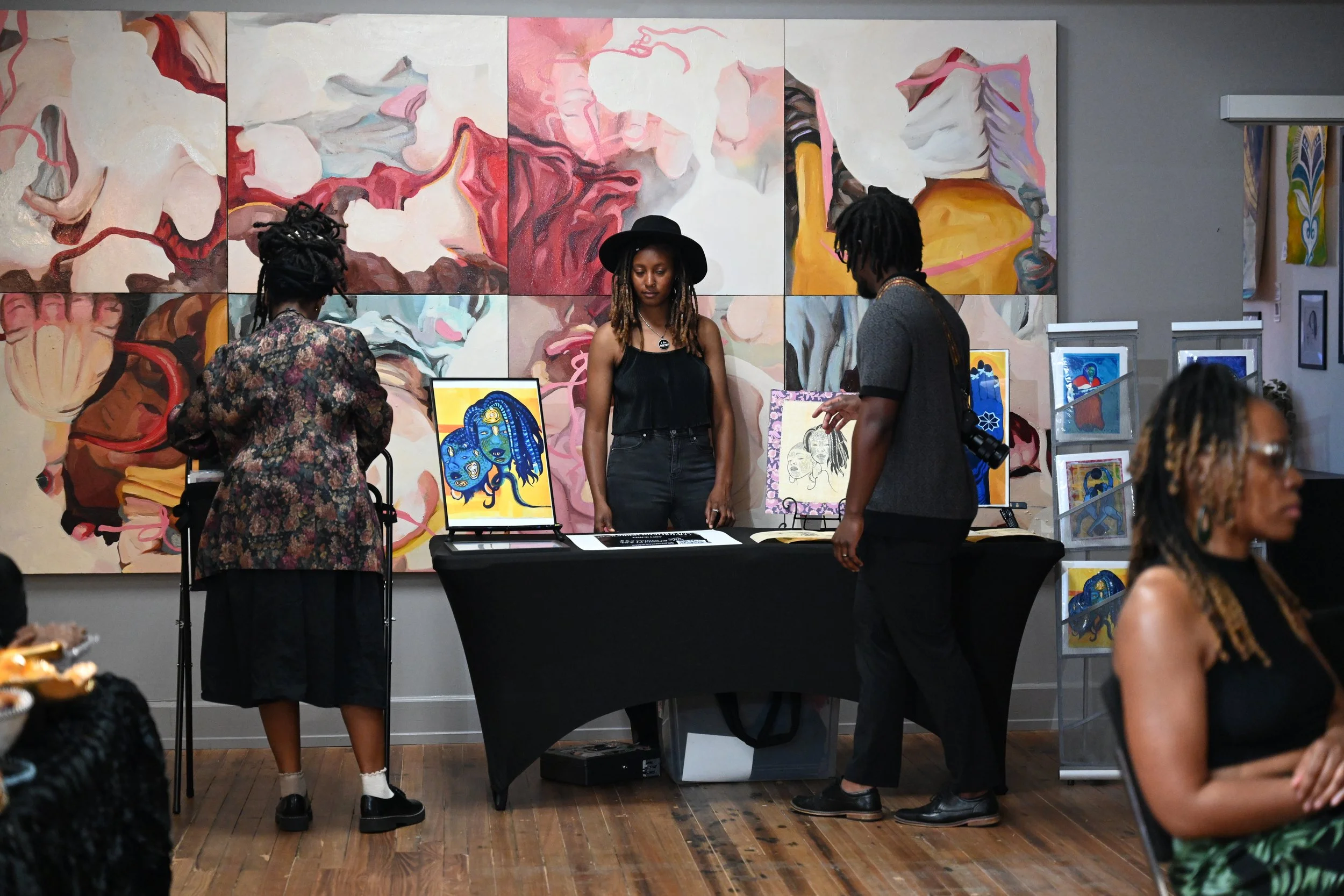 An art exhibition booth displaying vibrant paintings, including an abstract triptych with swirling colors, and smaller artwork featuring stylized portraits. Three people are engaging with the artwork, two women and one man, in a gallery with gray walls and wooden floors.