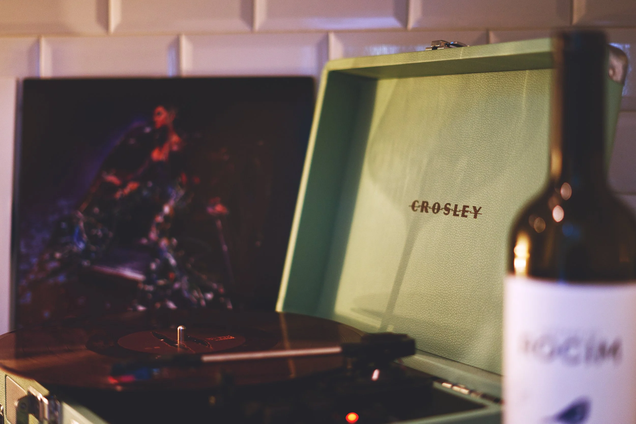 A turntable playing a record with a green Crosley speaker in the background, and a blurred colorful artwork or photograph is visible to the left.