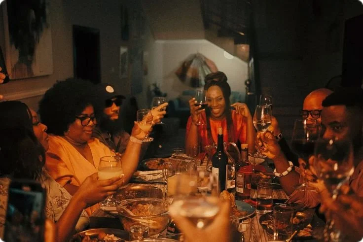 Group of people enjoying a dinner party, raising glasses for a toast, with food and drinks on the table in a dimly lit restaurant or home setting.