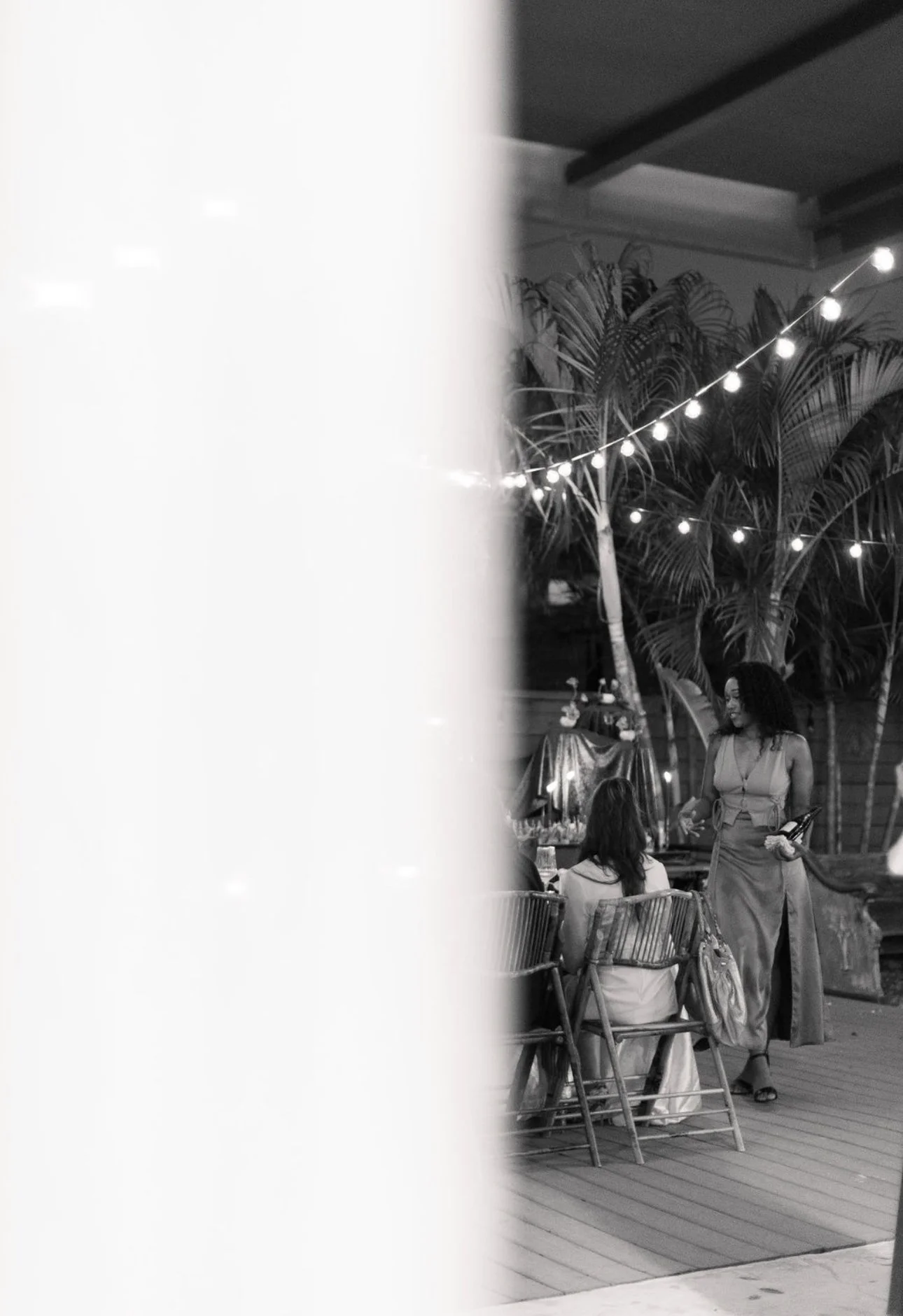 Two women dining outdoors at a restaurant or event with string lights, tropical plants, and a tent or canopy in the background.