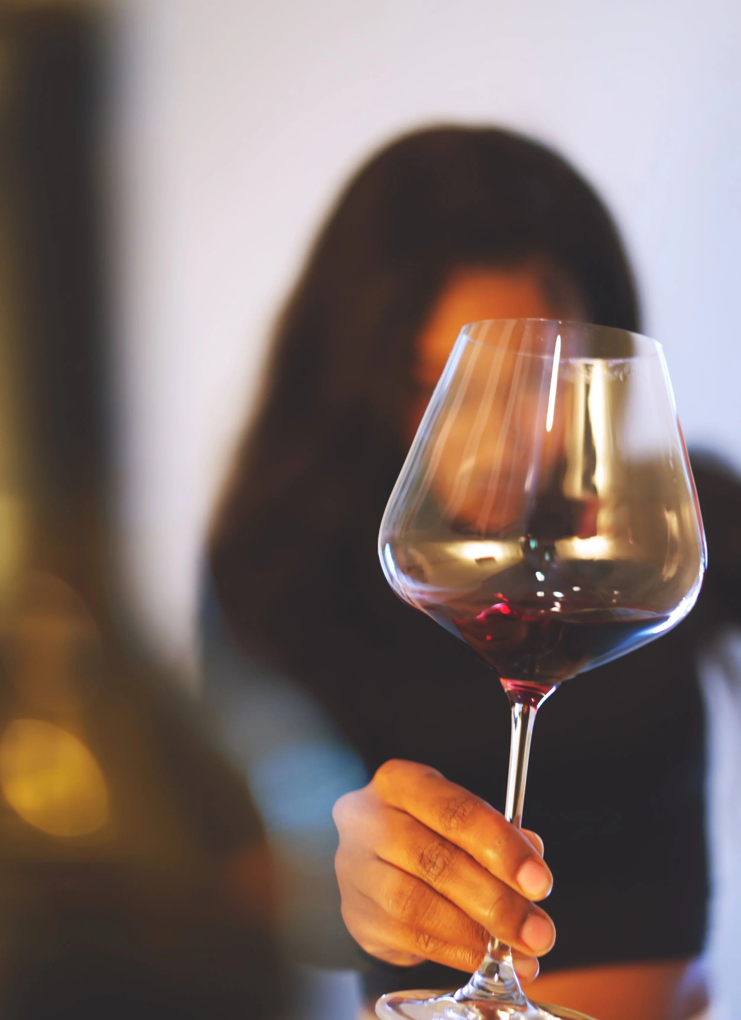 A person holding a glass of red wine with a blurred background of what appears to be a fireplace and a white wall.