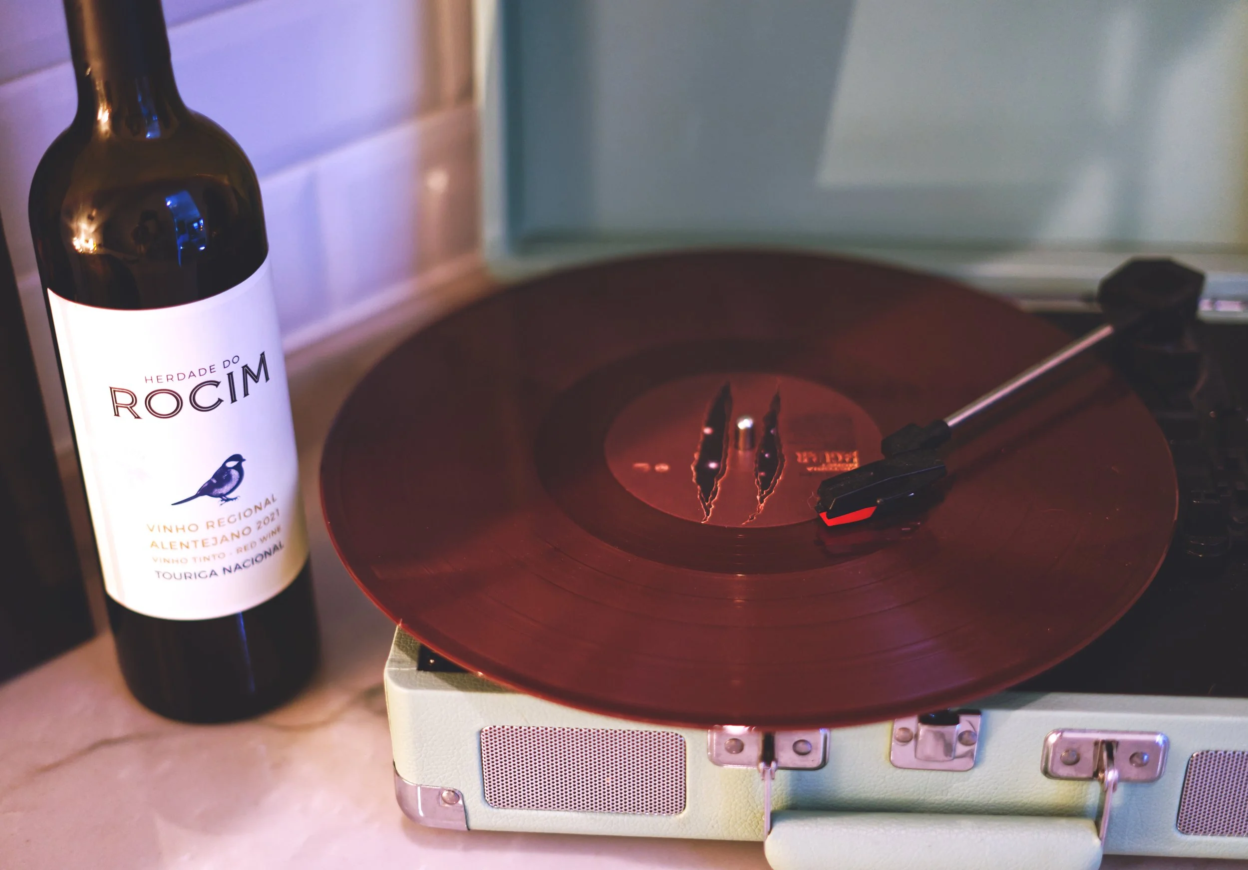 A bottle of Herdade do Rocim red wine with a blackbird logo and a red turntable playing records on a beige vintage-style portable speaker.