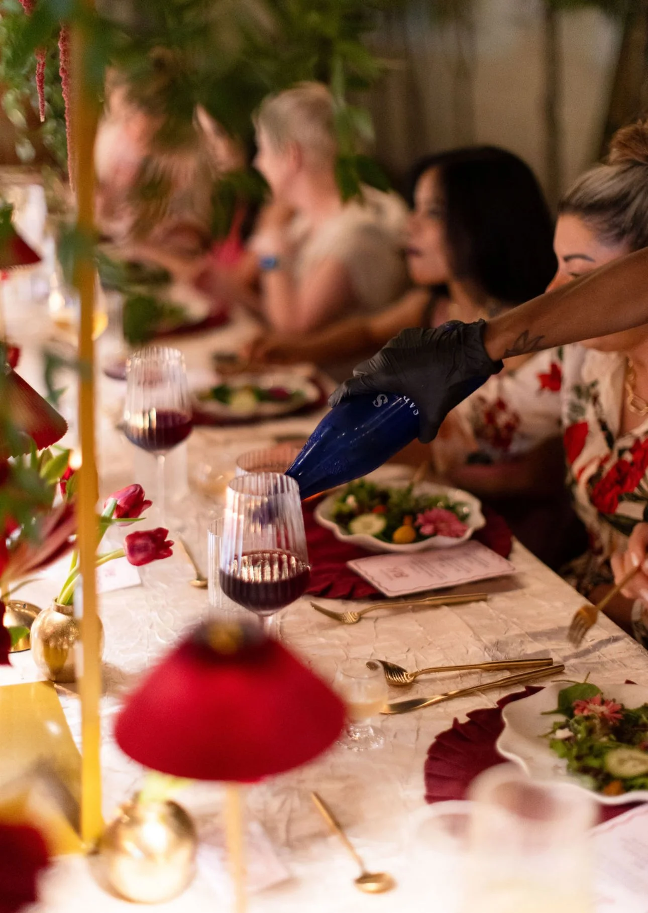 People seated at a festive dinner table with gold cutlery, wine glasses, and floral centerpieces, with one person pouring red wine into a glass.
