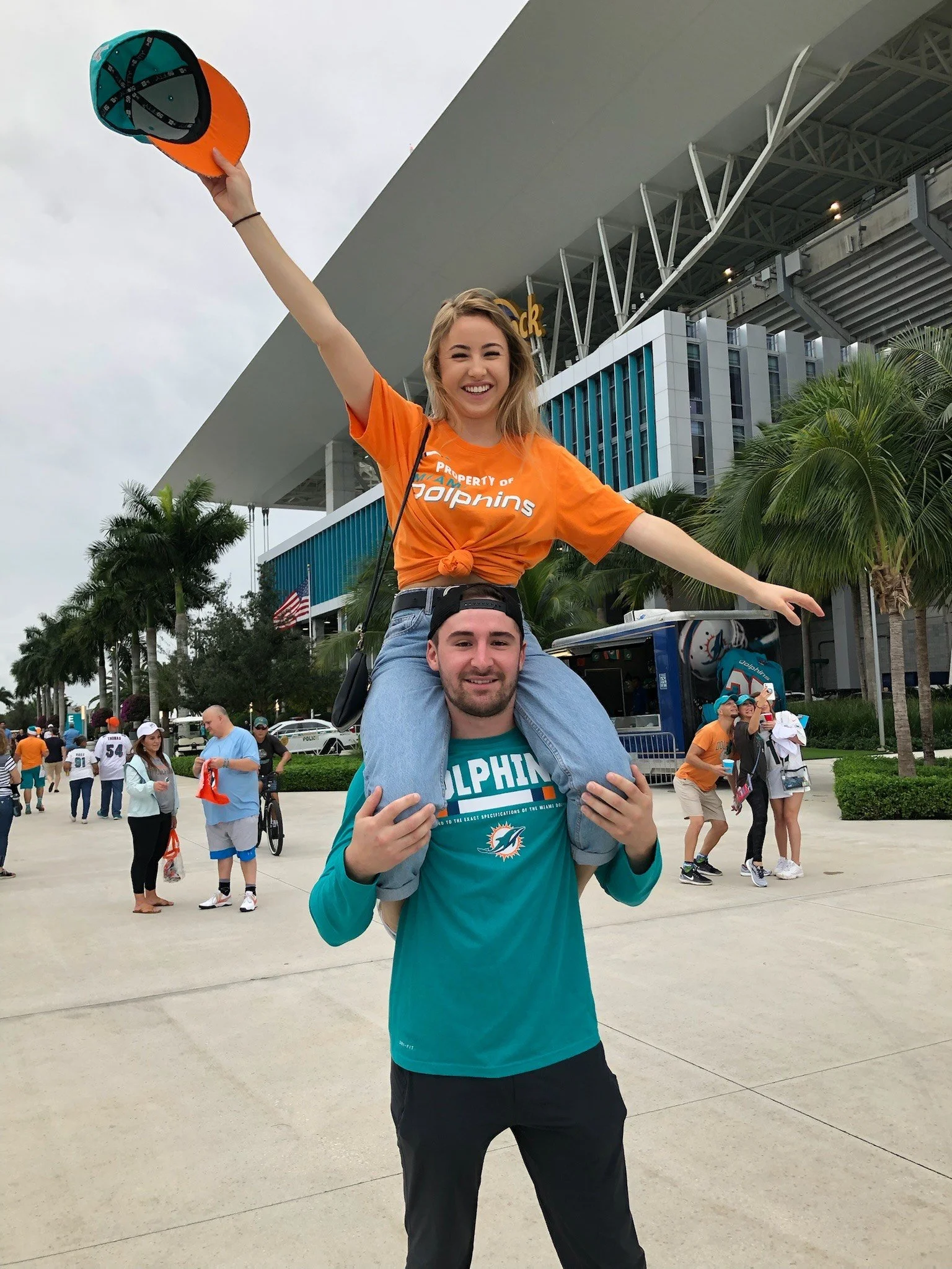 A young woman sitting on a young man's shoulders during a celebration outside a stadium. The woman is smiling, holding a cap in the air, and wearing an orange T-shirt. The man is wearing a teal Miami Dolphins shirt, smiling, and holding the woman's l