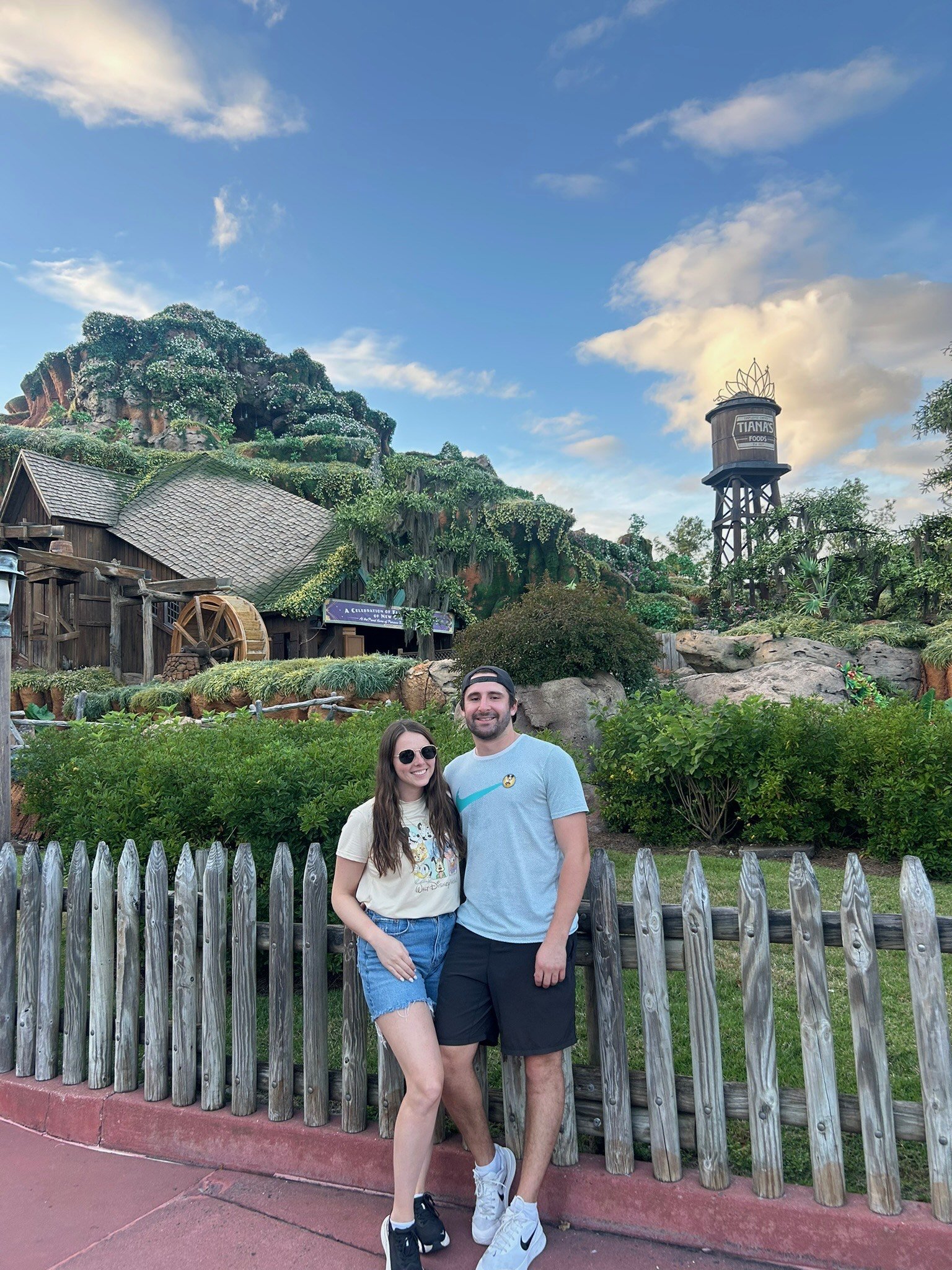 A smiling young woman and a smiling young man standing close together outdoors behind a wooden fence, with greenery and a themed building resembling a fairytale castle in the background, along with a water tower labeled 'Tianka's Foods', under a part