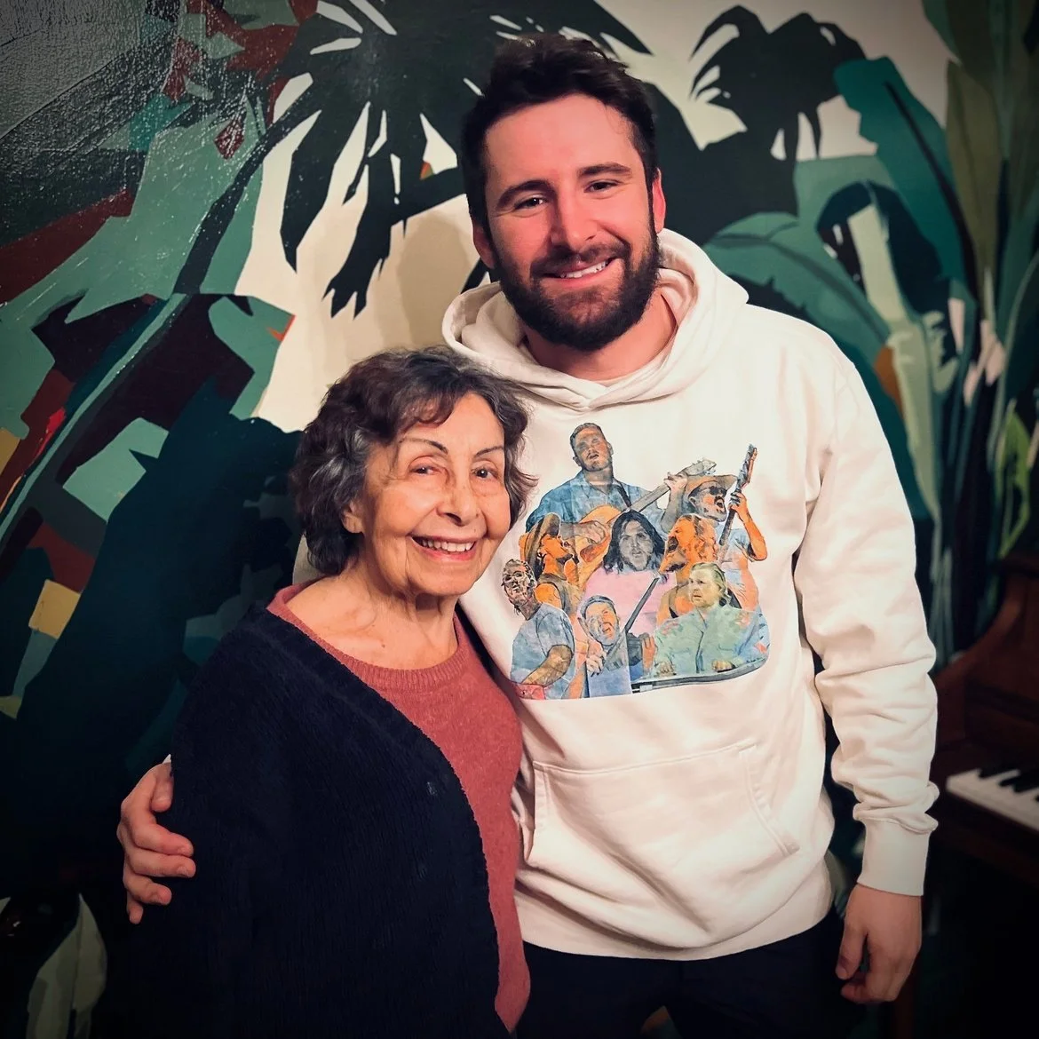 A smiling older woman with curly gray hair standing next to a young man with a beard, both posing indoors in front of a colorful jungle-themed mural. The young man is wearing a white hoodie with a printed design of various musicians.
