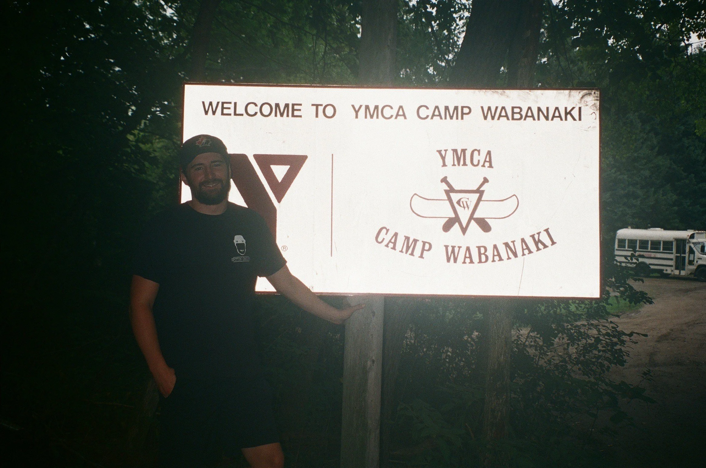 A man with a beard and dark clothing standing outdoors next to a white signboard that reads "Welcome to YMCA Camp Wabanaki" with a YMCA logo on it, surrounded by trees and a truck in the background.