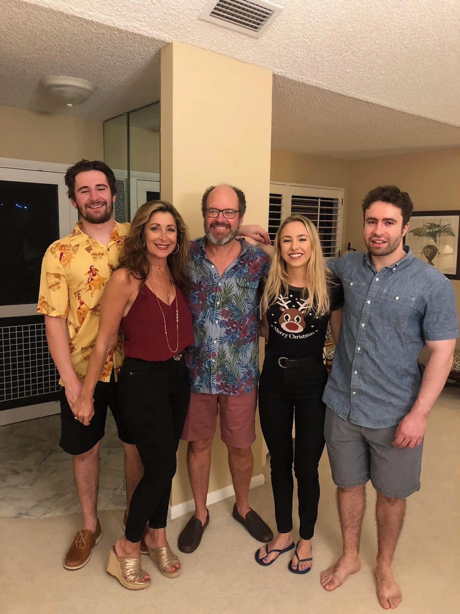 A family standing together in a room, smiling, wearing casual clothing and sandals or barefoot.