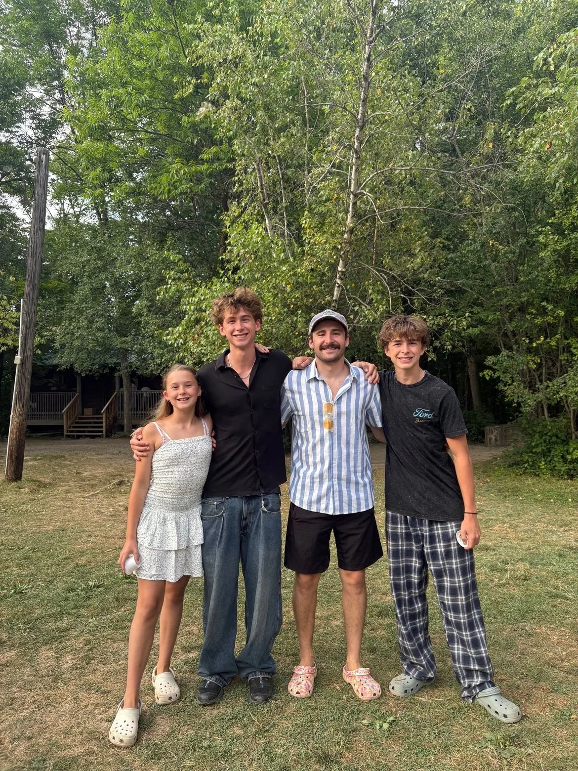 Four smiling people standing outdoors in front of green trees, with two young guys in the middle, a young girl on the left, and a teenage boy on the right. They are casually dressed and have their arms around each other.